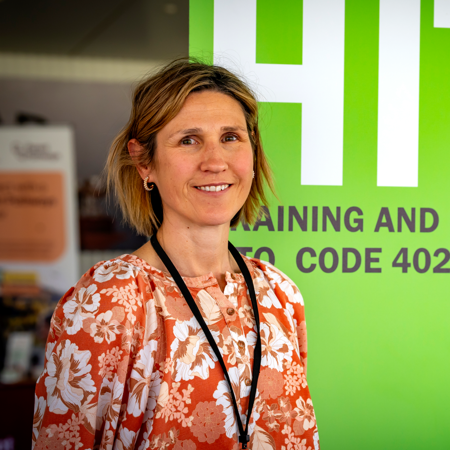 Close-up of a woman smiling, with a bright green sign in the background that has large white and black text.