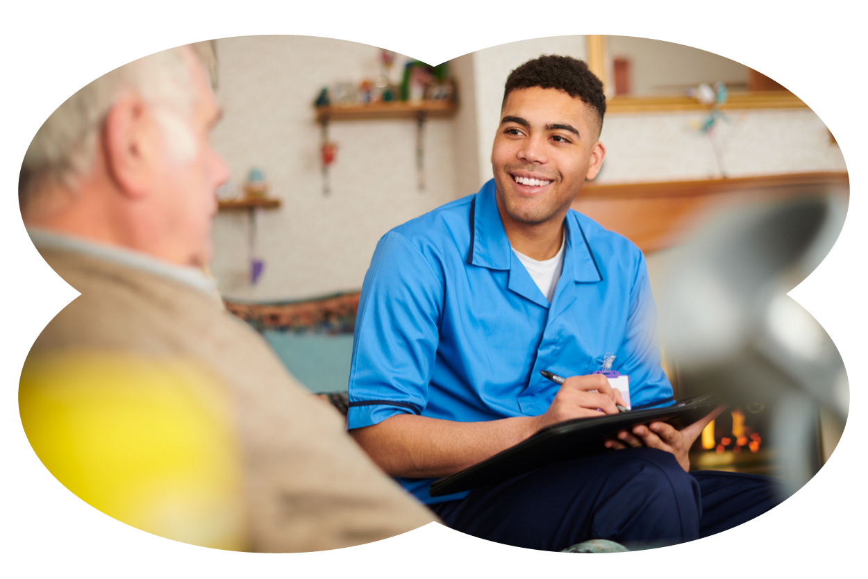 A young man in a blue uniform smiling and holding a clipboard while talking to an elderly man in a home setting.