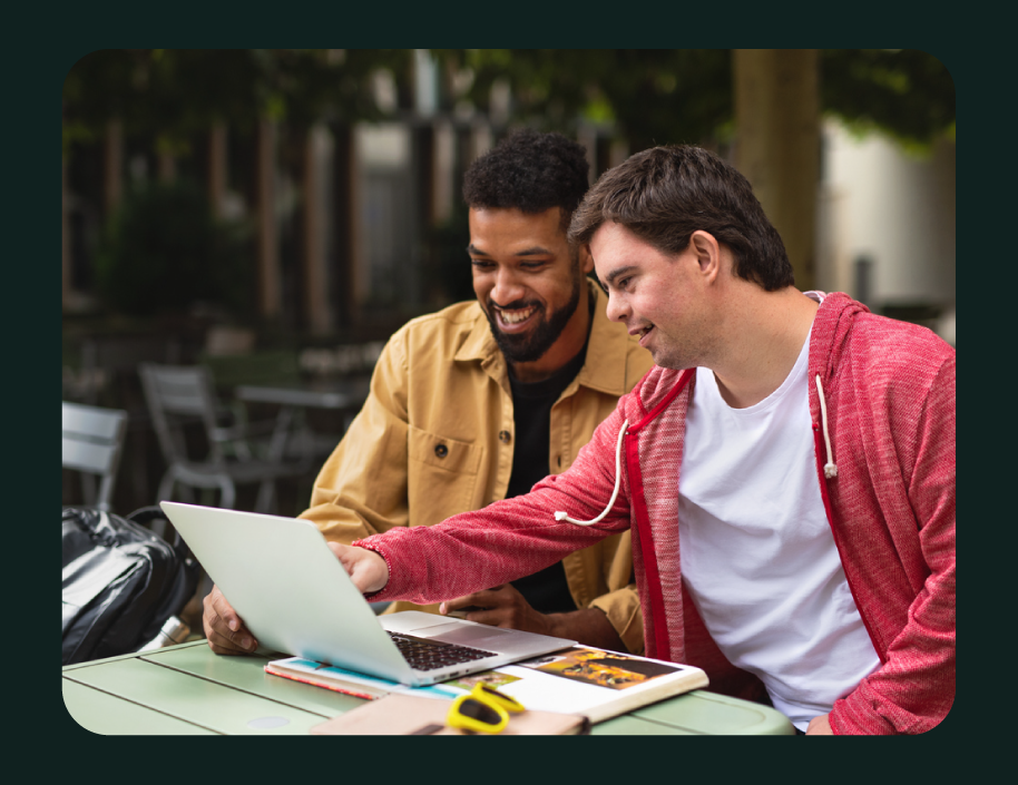 Two young men sitting at an outdoor table looking at a laptop, smiling and engaged in conversation, with books and a backpack on the table.