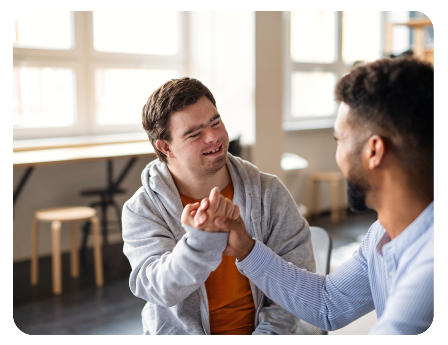 Two men are smiling and engaging in a friendly handshake or bump in a bright room with large windows and minimal furniture.