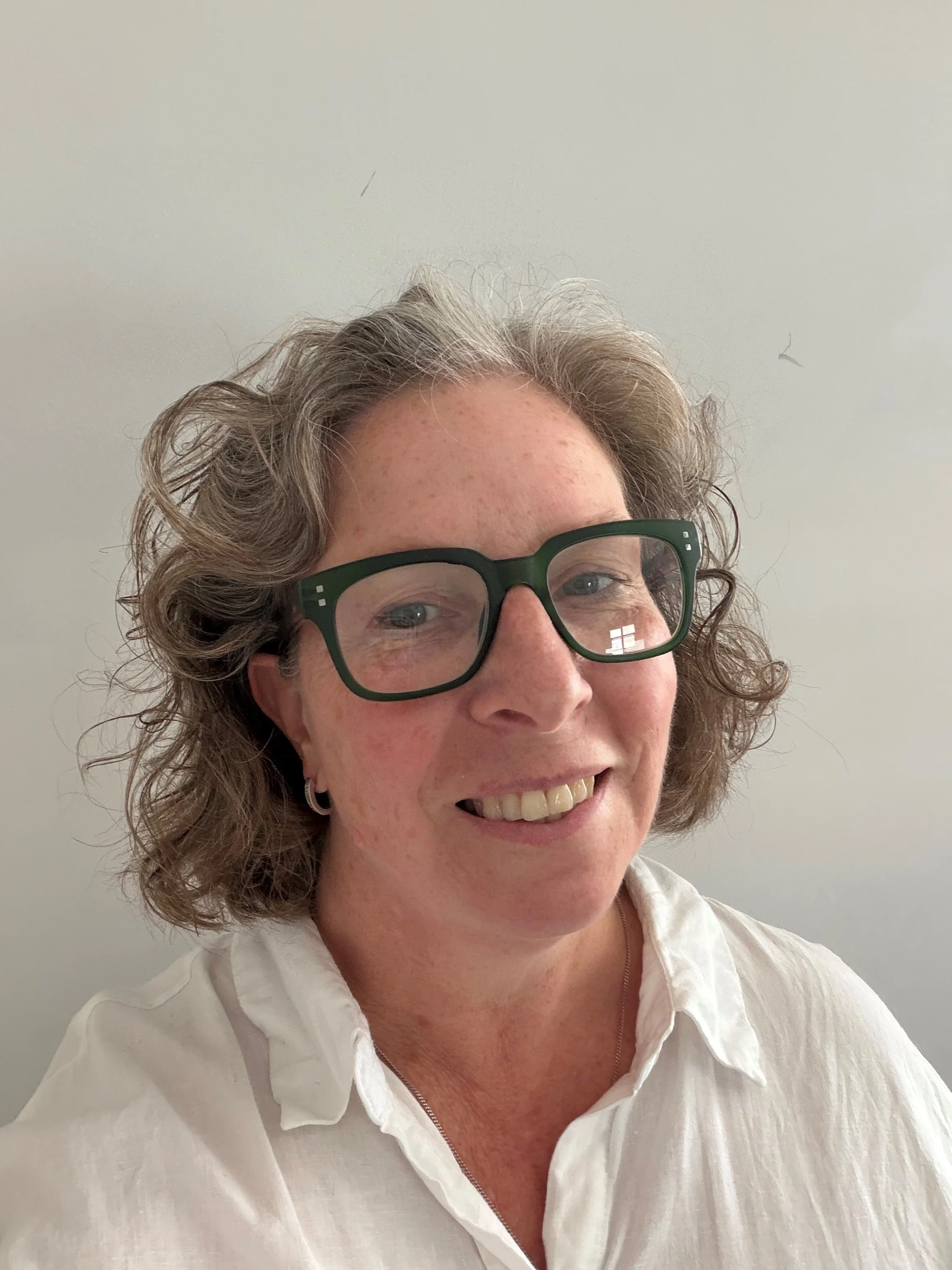 A woman with curly hair wearing glasses and a white shirt, smiling in front of a plain light-colored background.