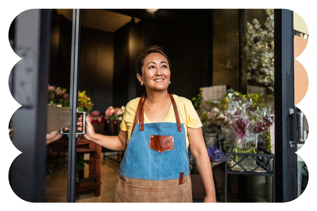 A woman in a yellow shirt and apron standing inside a flower shop, holding a door open, with flowers visible in the background.