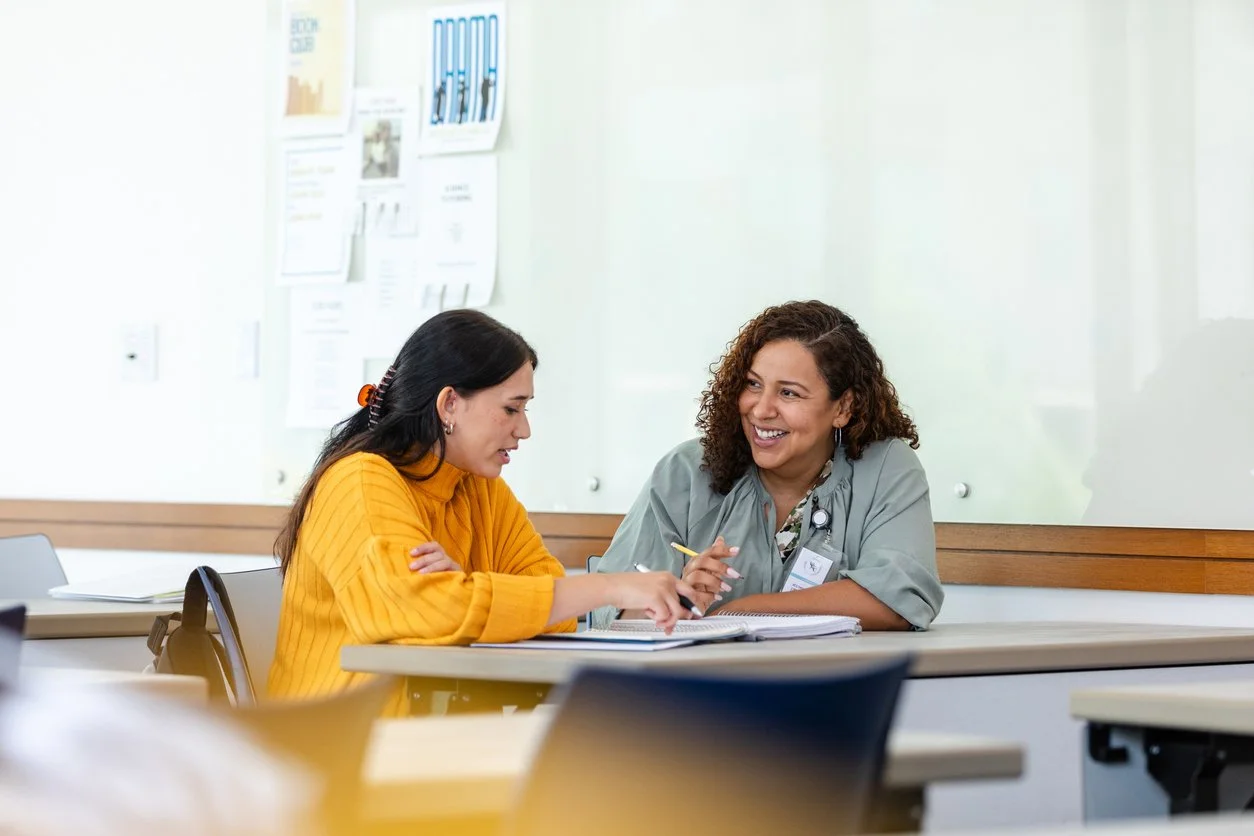 Two women sitting at a table in a classroom or training room, engaged in conversation. One woman is wearing a yellow sweater and the other is wearing a gray shirt, smiling and talking.