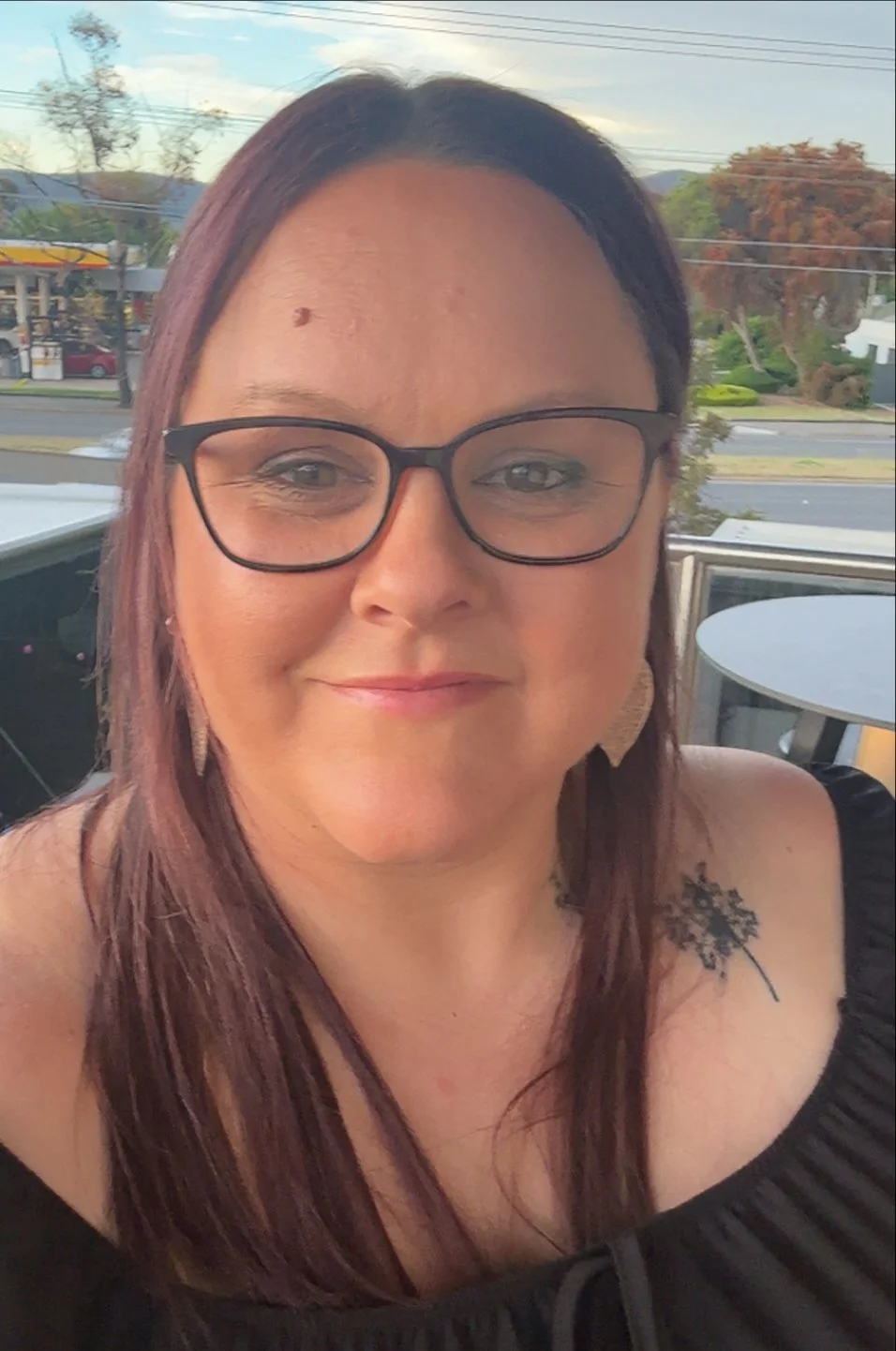 A woman with glasses and long brown hair smiling outdoors, wearing a black off-shoulder top with a floral tattoo on her shoulder, in front of a window with a street and trees in the background.