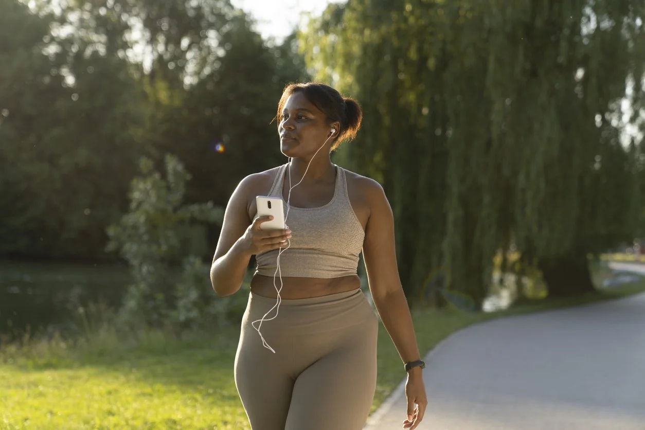 Woman walking outdoors in athletic wear, holding a smartphone and listening to music with earphones, on a sunny day near a park or river.