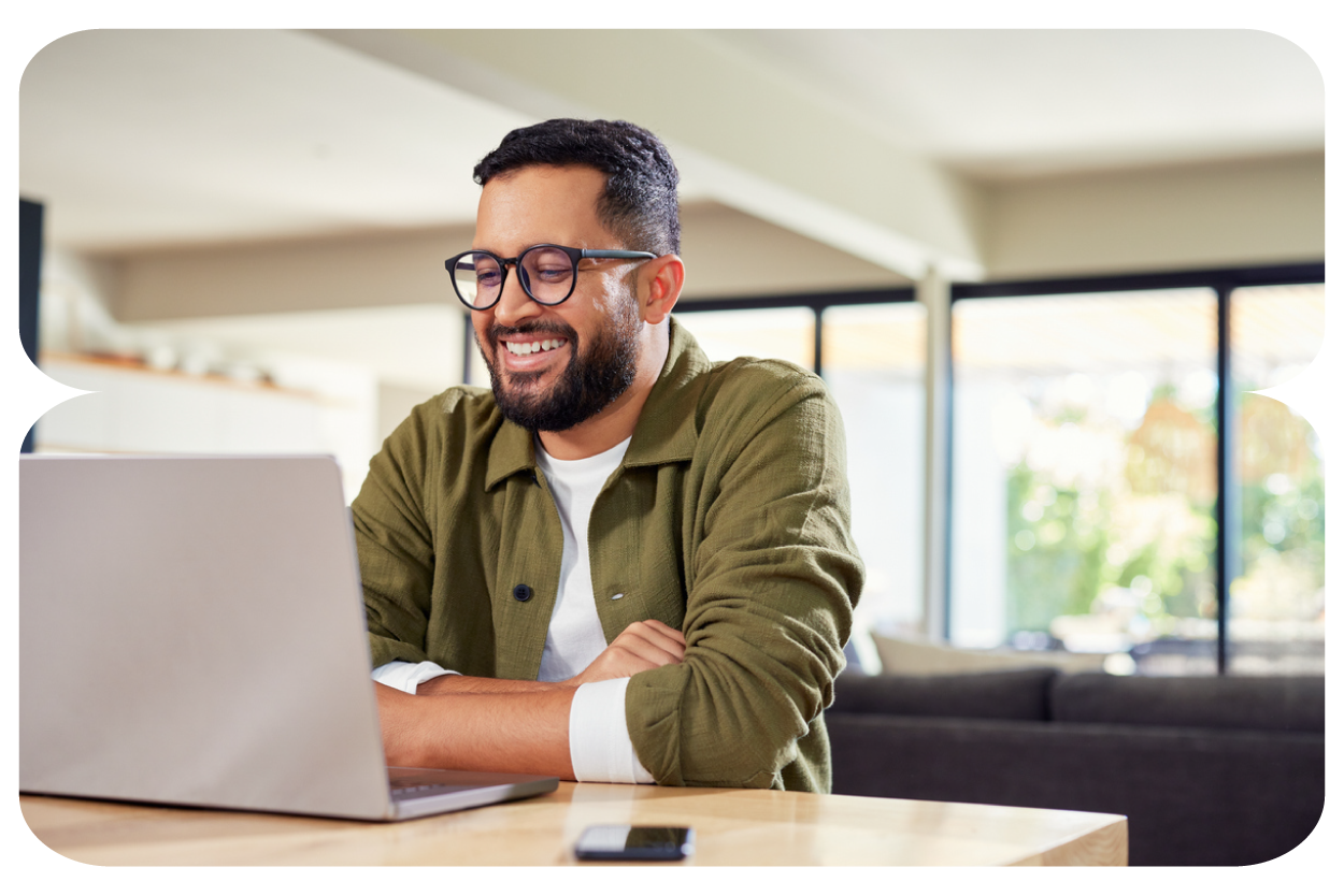 A smiling man with glasses and a beard working on a laptop at a wooden table in a bright, modern room with large windows.