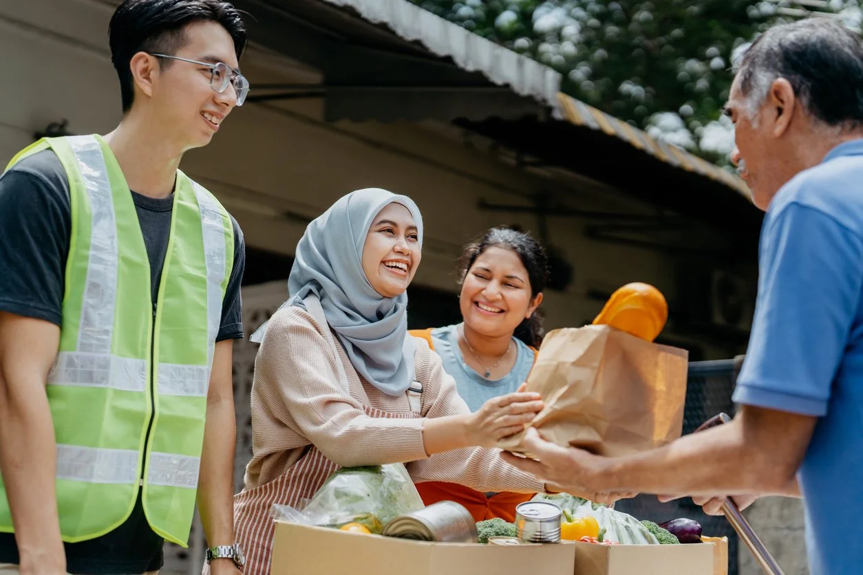 People volunteering at a food drive, handing a paper bag of groceries to an elderly man, with groceries on the table.