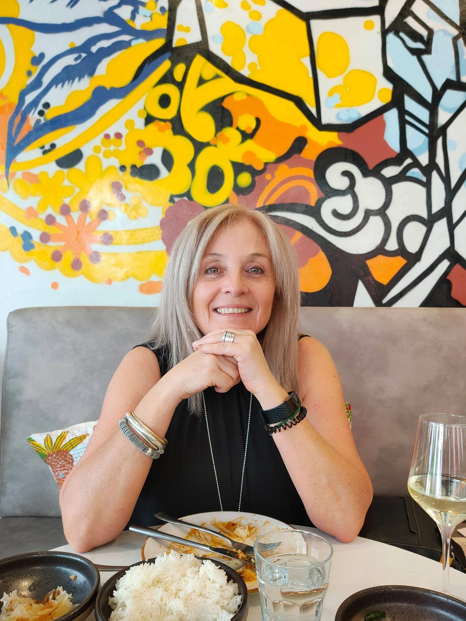 A woman with blonde hair smiling while sitting at a restaurant table with food, wine glass, and decorative background.
