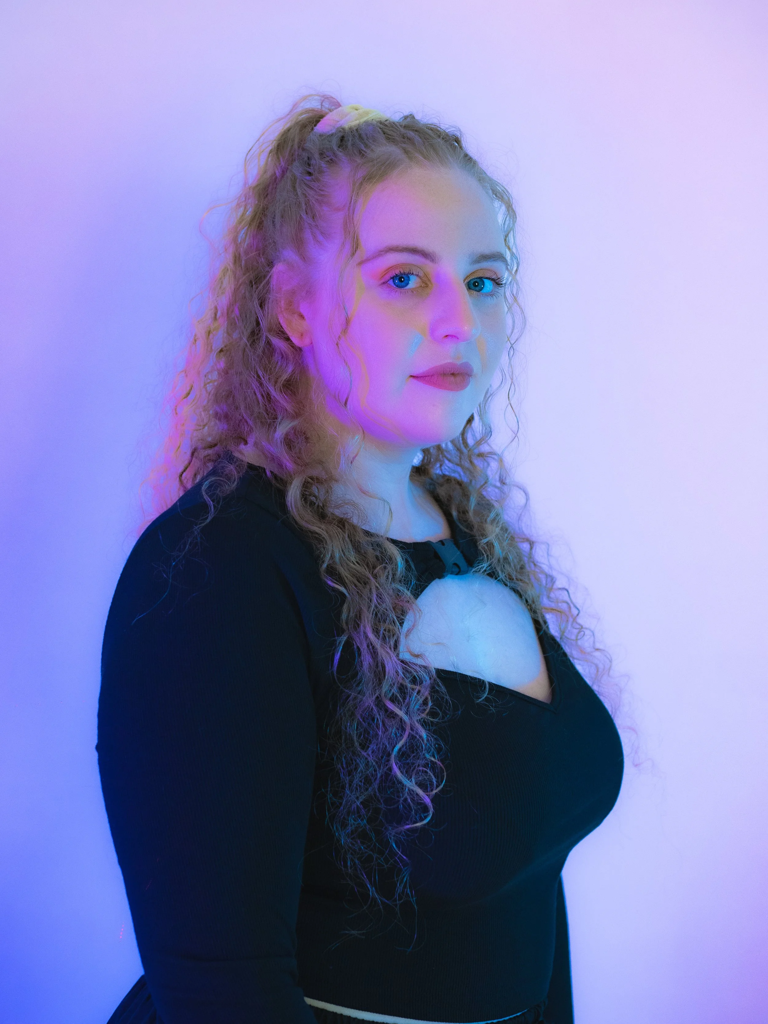 A woman with curly blonde hair tied in a ponytail, wearing a black top, stands against a plain white wall illuminated with purple and pink lighting.