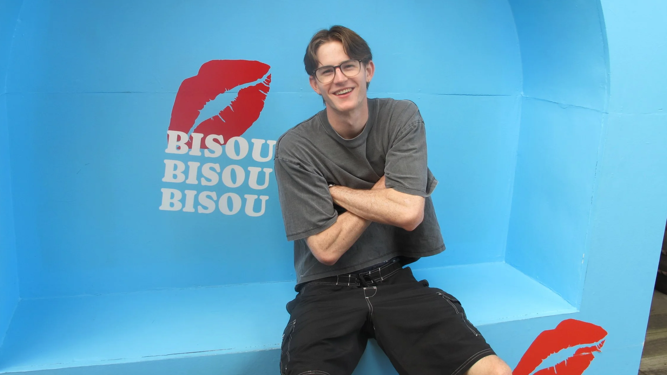 Young man with glasses smiling, sitting cross-armed in a blue photo booth with a red lipstick kiss mark and the repeated word 'BISOU' in white letters.