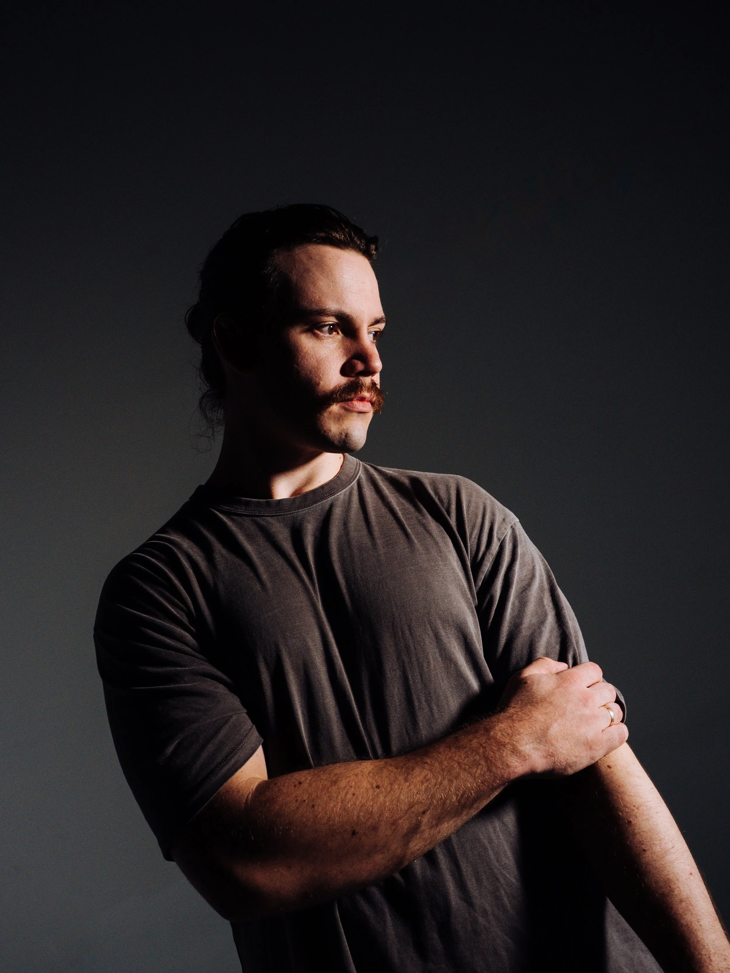 A man with dark hair, a mustache, and a beard, wearing a brown t-shirt, standing against a dark gradient background, with his arms crossed, gazing to the left.