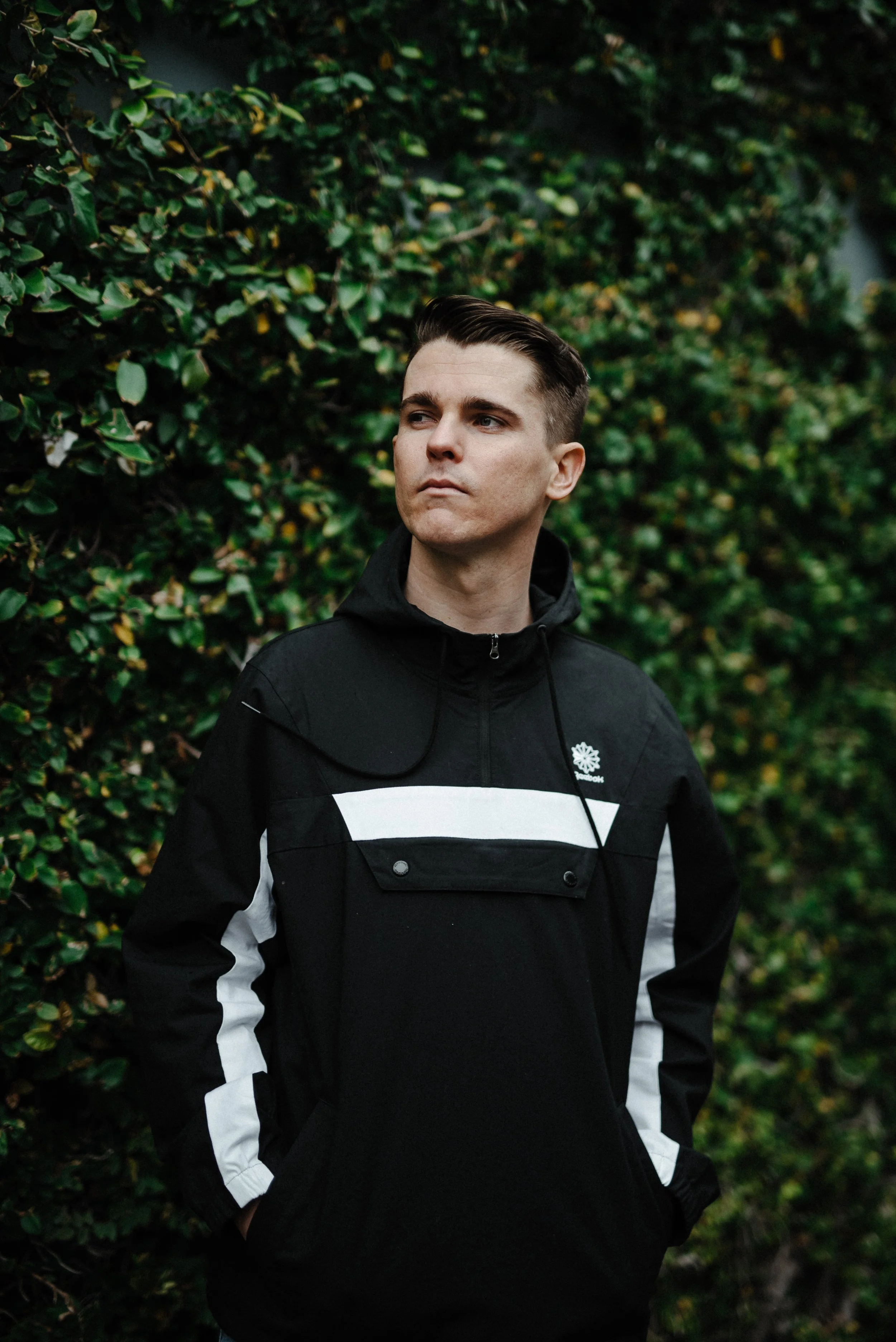 A young man with short dark hair and pale skin stands outdoors in front of a dense green hedge, wearing a black Reebok jacket with white accents and a logo on the chest, looking slightly to his right with a serious expression.