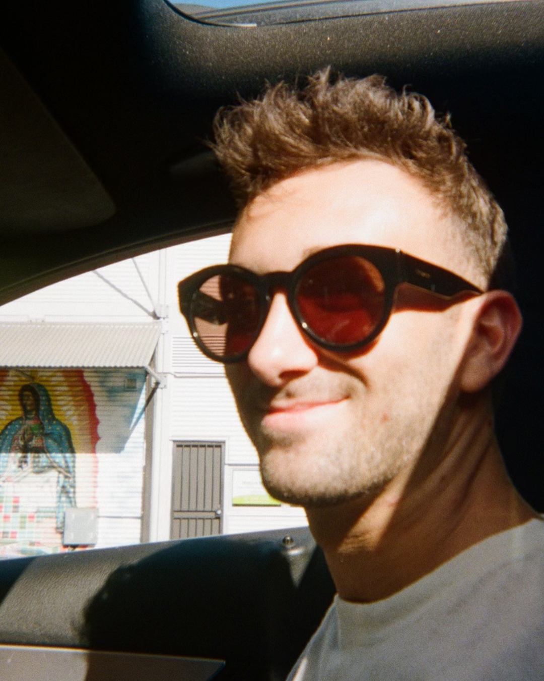 A young man with curly hair wearing large round sunglasses, sitting in a car with sunlight on his face.