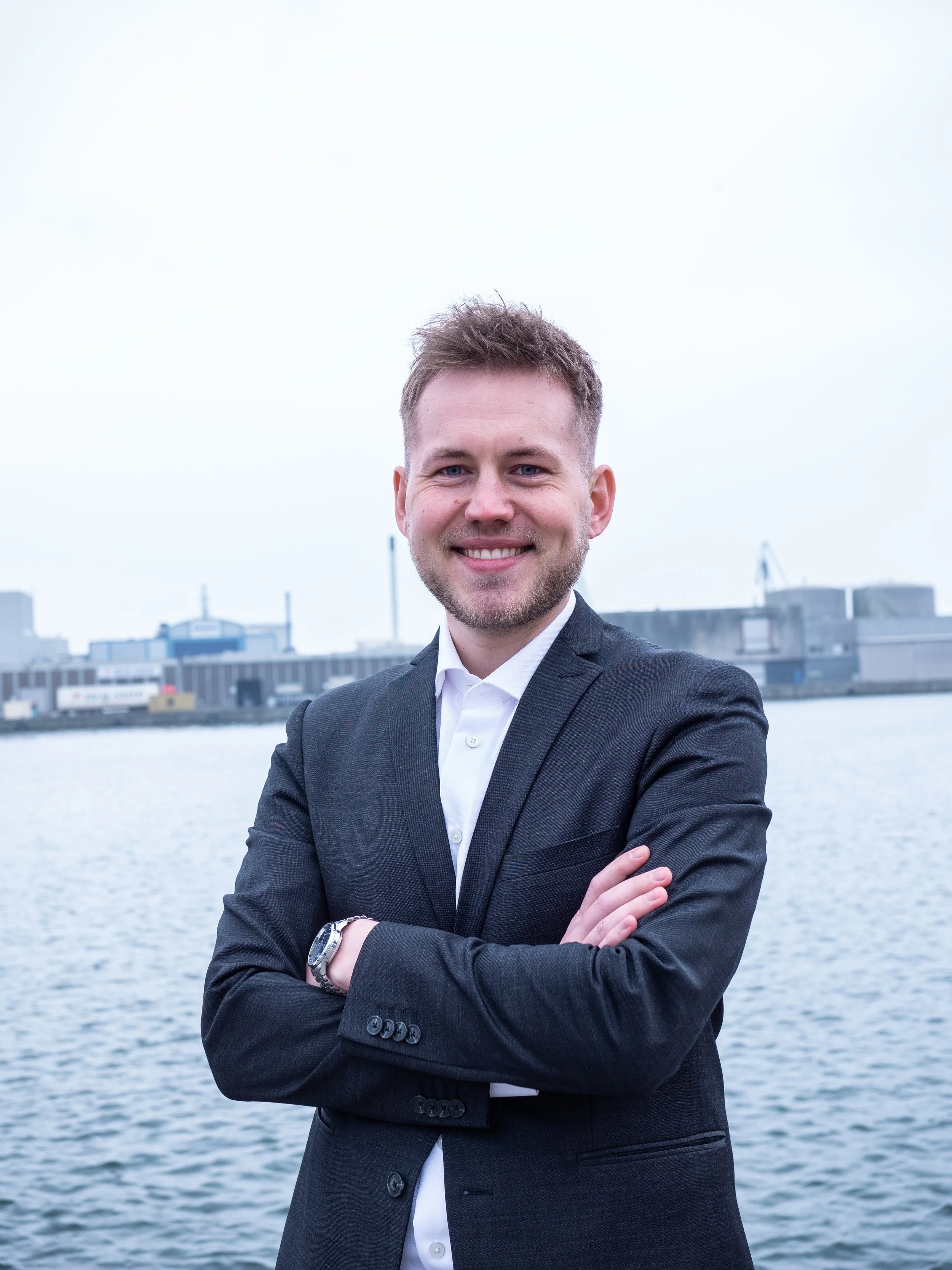 A man in a dark suit and white shirt standing outdoors by a body of water, with industrial buildings in the background, smiling with arms crossed.
