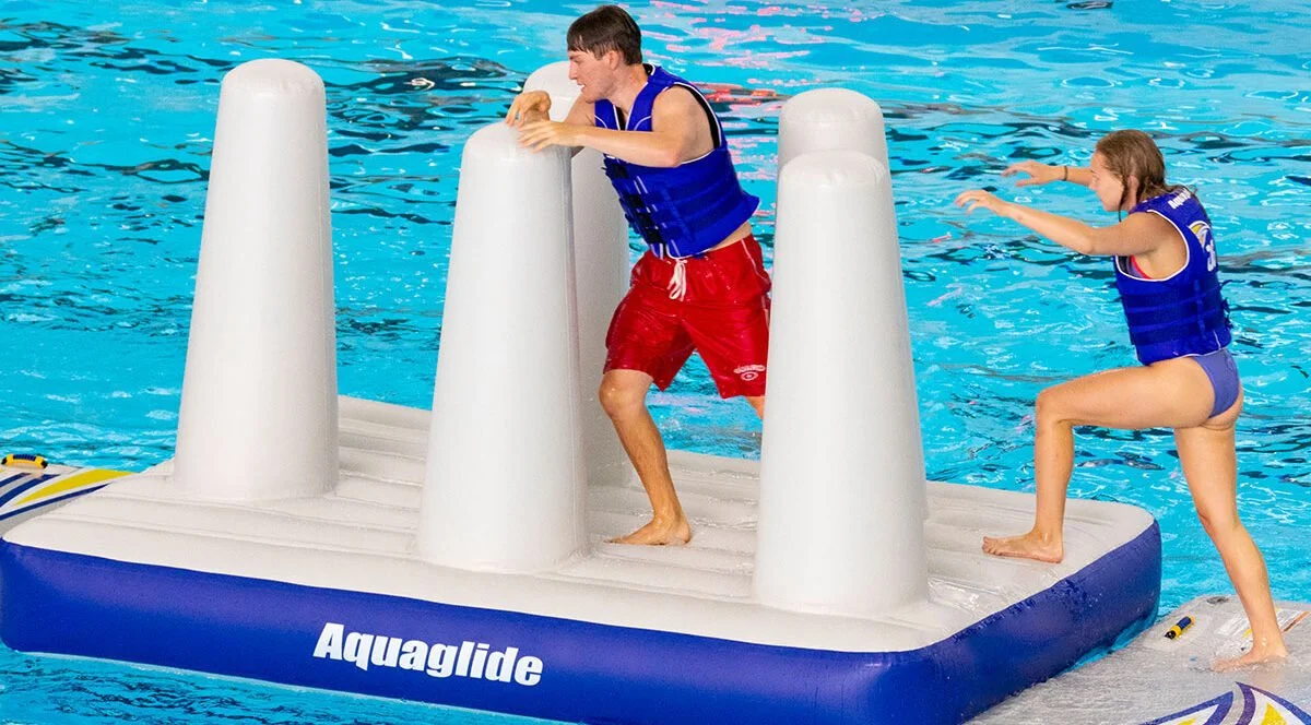 Two children on an Aquaglide inflatable platform in a swimming pool, one helping the other to stay balanced.