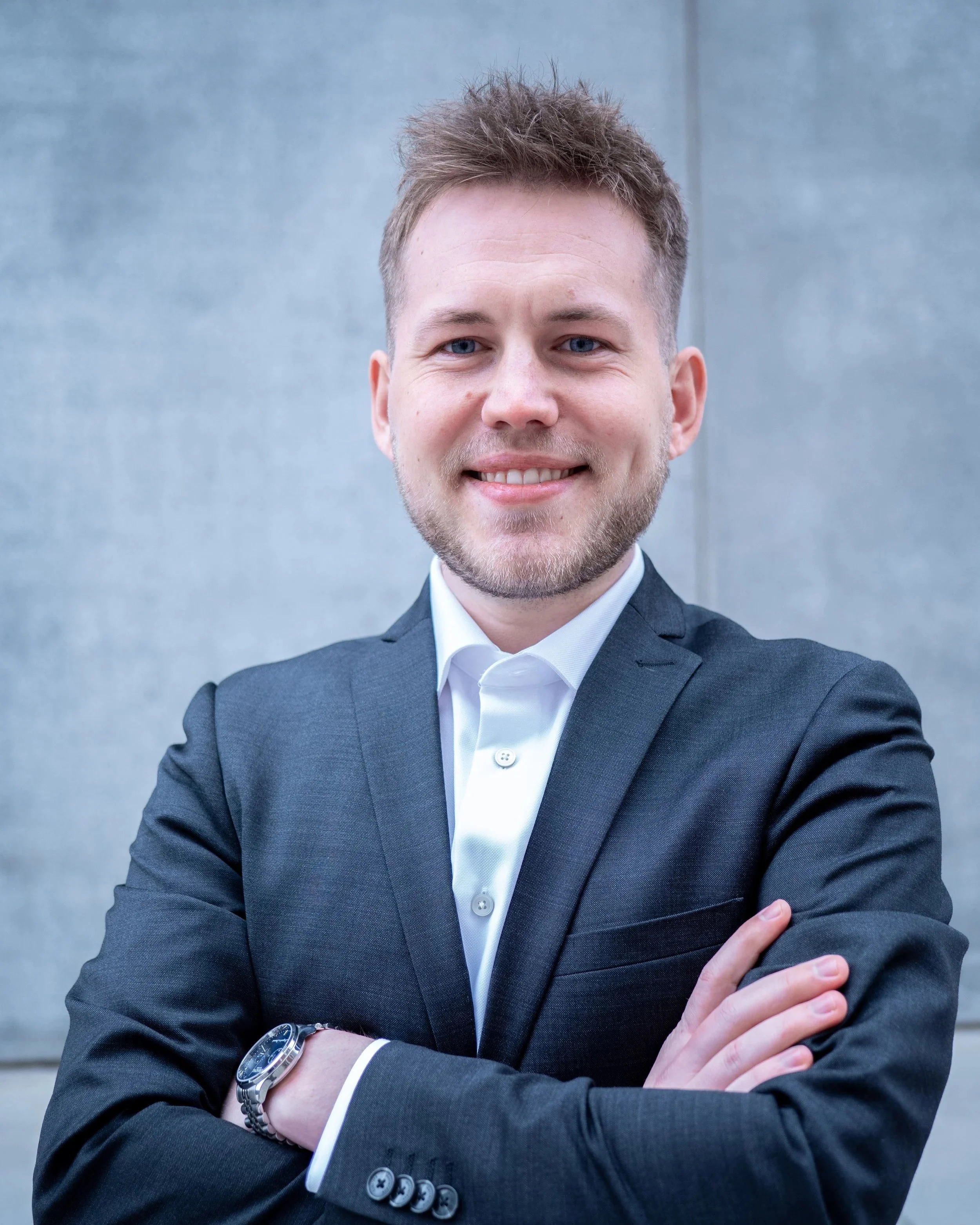 Close-up of a young man with light brown hair, blue eyes, and light facial hair, wearing a dark gray suit, white shirt, and silver wristwatch, standing outdoors with a gray concrete wall in the background.