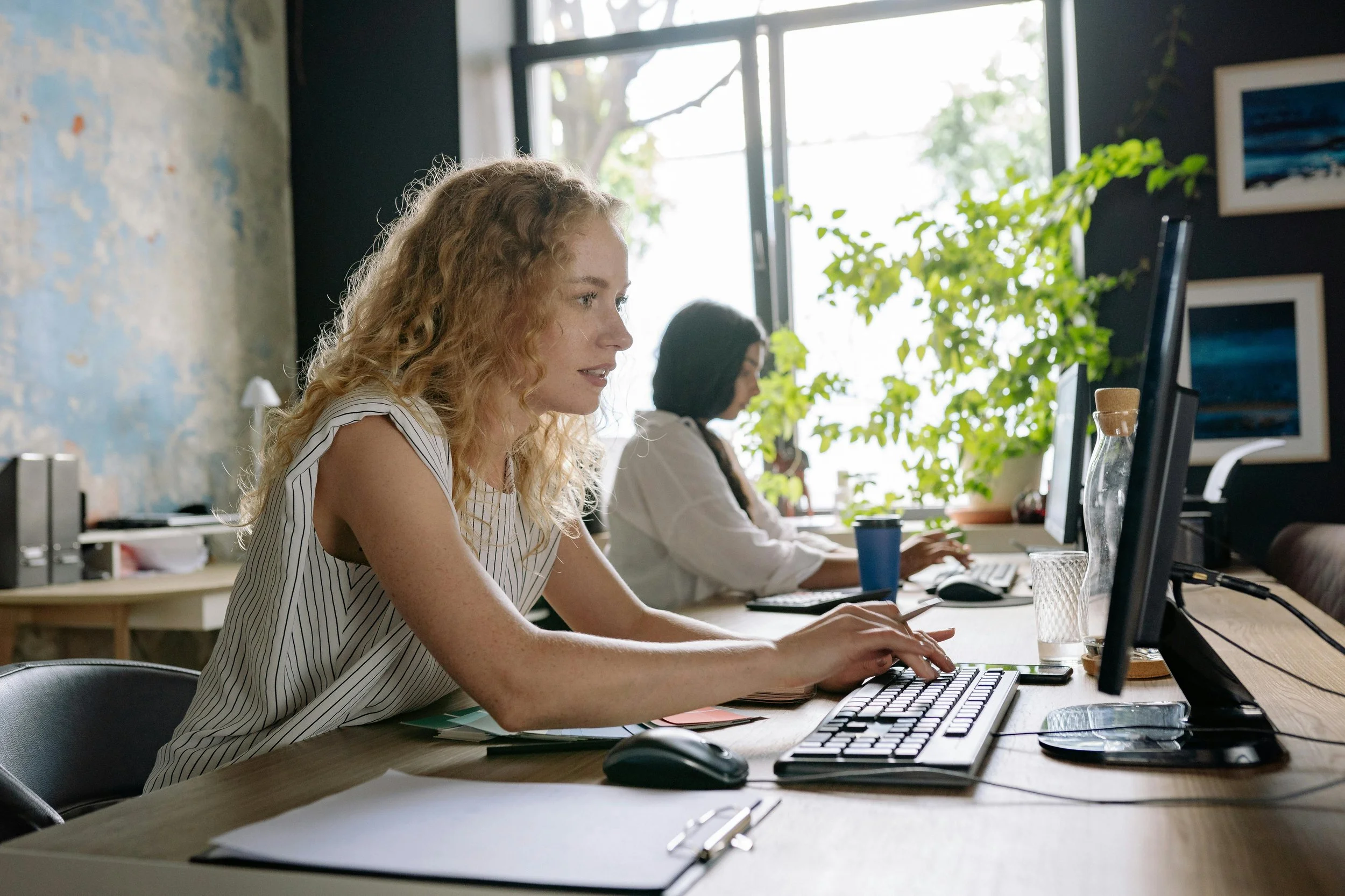 Two women working at desks in a bright office with large windows, plants, and artwork on the walls.
