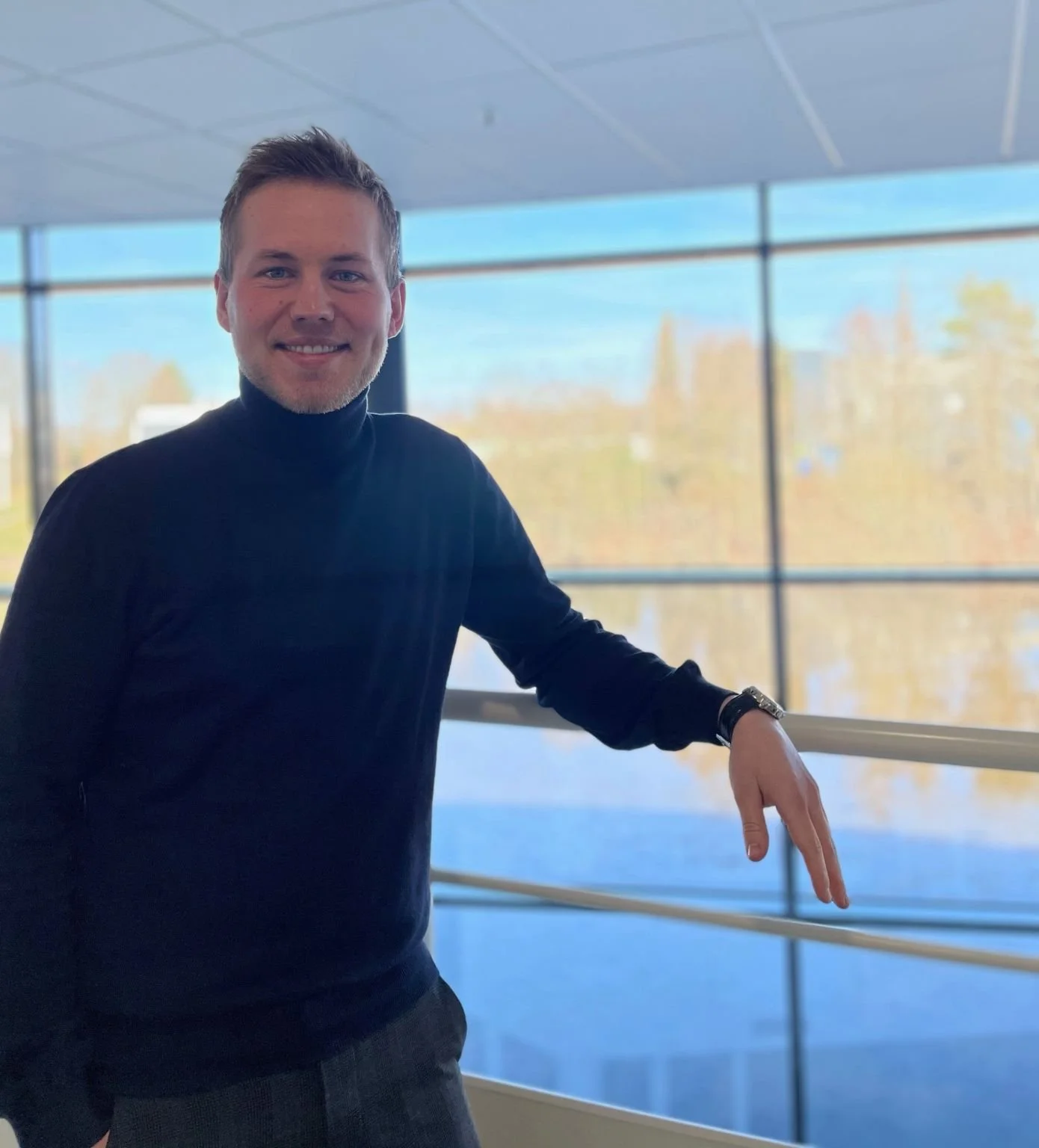 A young man with short hair and a beard, wearing a black turtleneck sweater, standing inside a building with large windows showing trees and a blue sky outside. He is smiling and resting one arm on railing.