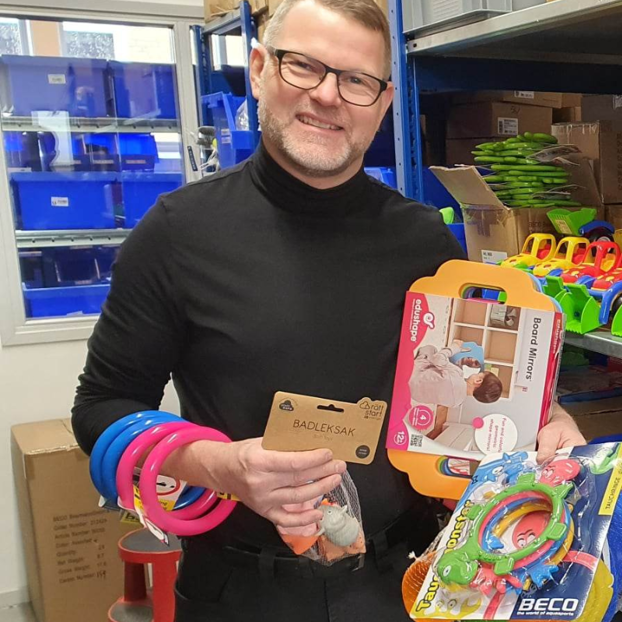 Man shopping for children's toys in a store, holding colorful bracelets, a drawing board, and a toy set with oscillating rings, with shelves and boxes in the background.