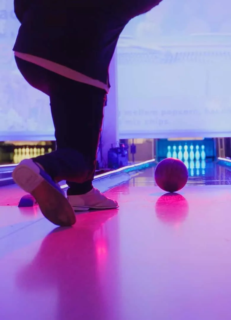 Close-up of a person crouching near a bowling ball on a bowling alley illuminated with colorful neon lights.