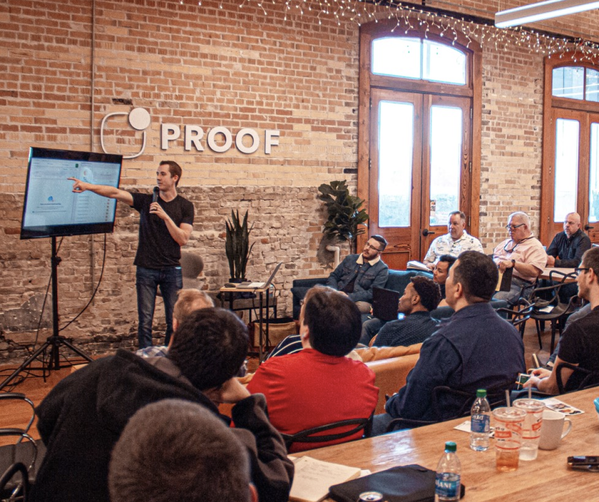 A presentation or meeting taking place in a room with brick walls, large windows, and hanging string lights. A man is standing in front of a screen, pointing at it while holding a microphone, addressing an audience seated around tables.