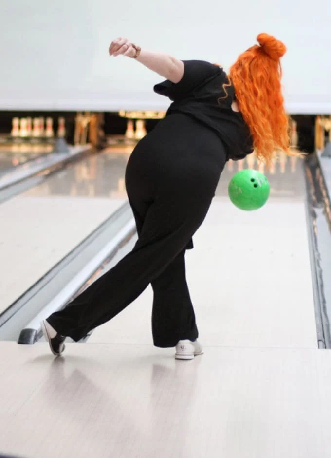 A woman with red curly hair wearing black clothing, in a bowling alley, throwing a green bowling ball down the lane.