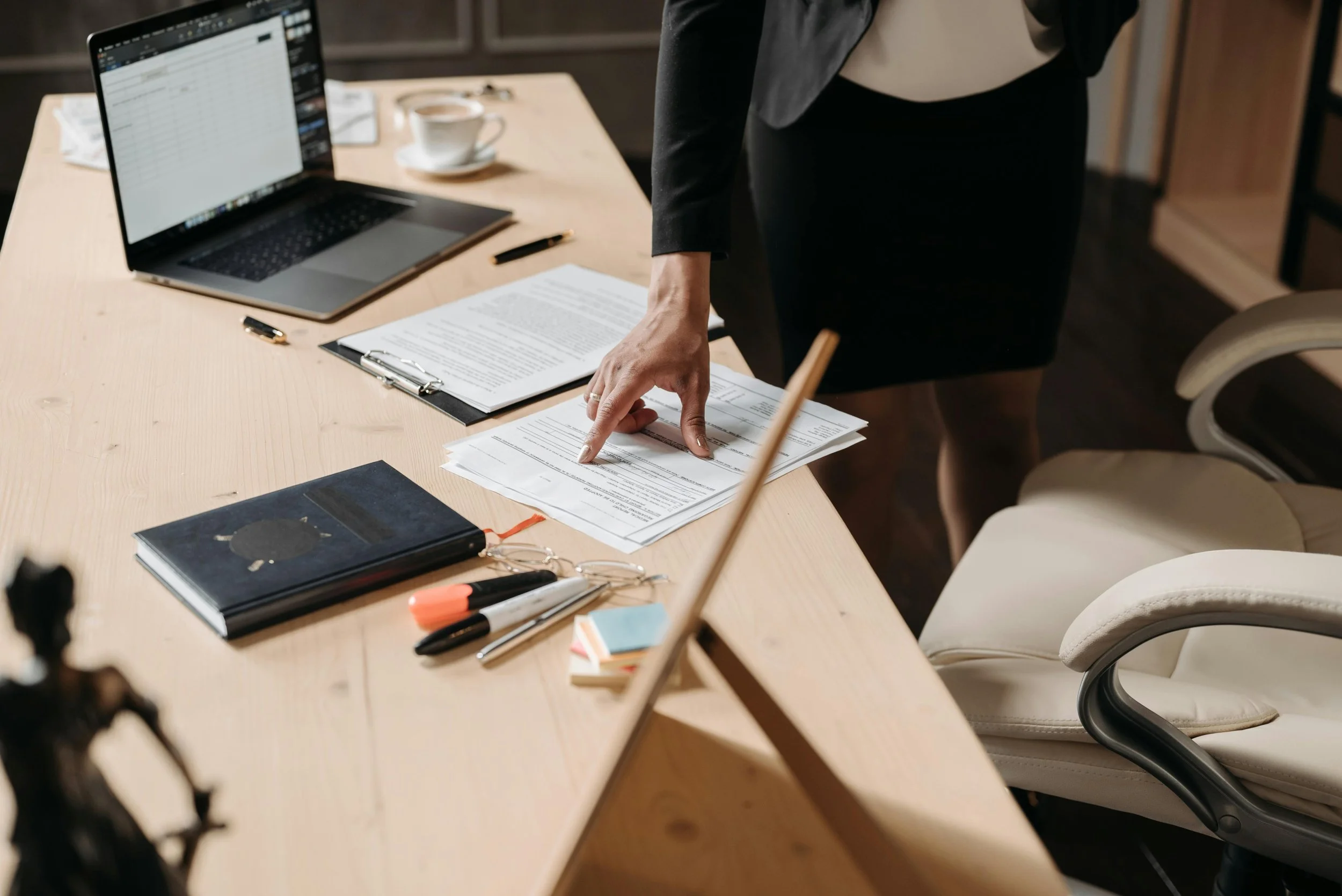 Person pointing at documents on a desk with a laptop, coffee cup, and office supplies.