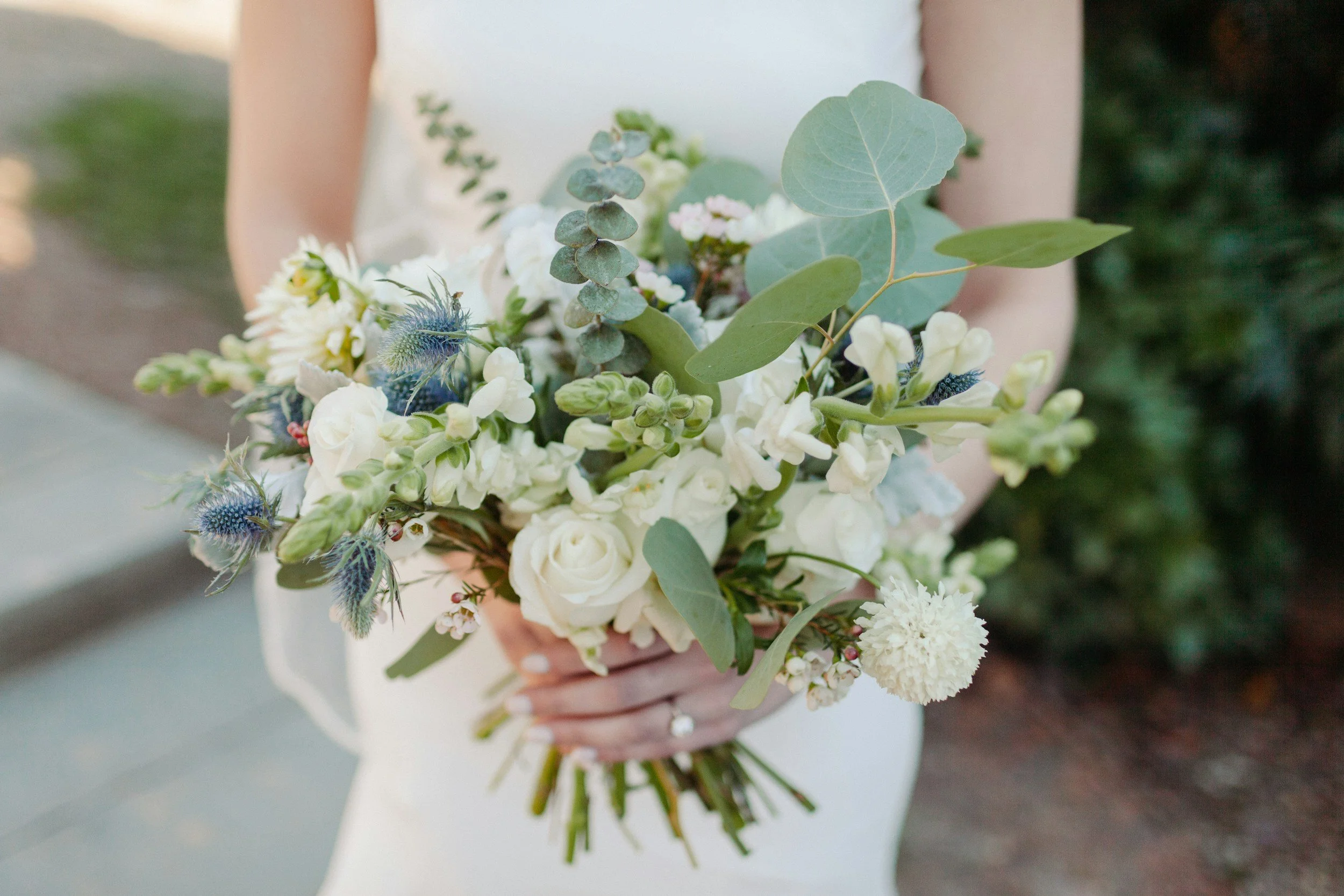 A person in a wedding dress holding a bouquet of white and green flowers with some blue accents and eucalyptus leaves.