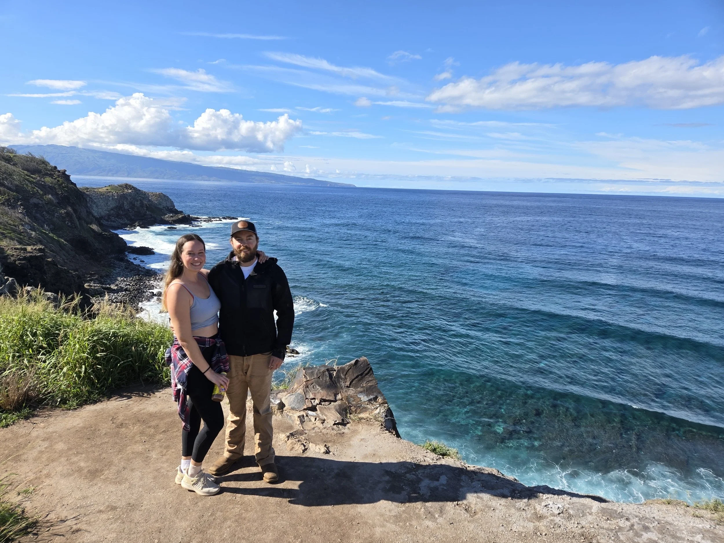 A smiling couple stands on a dirt trail by the ocean, with green grass and rocks in the foreground. The woman is wearing a gray crop top, black leggings, and sneakers, and the man is wearing a black jacket, tan pants, and a cap. They are near a rocky cliff edge with the ocean behind them, under a partly cloudy sky.