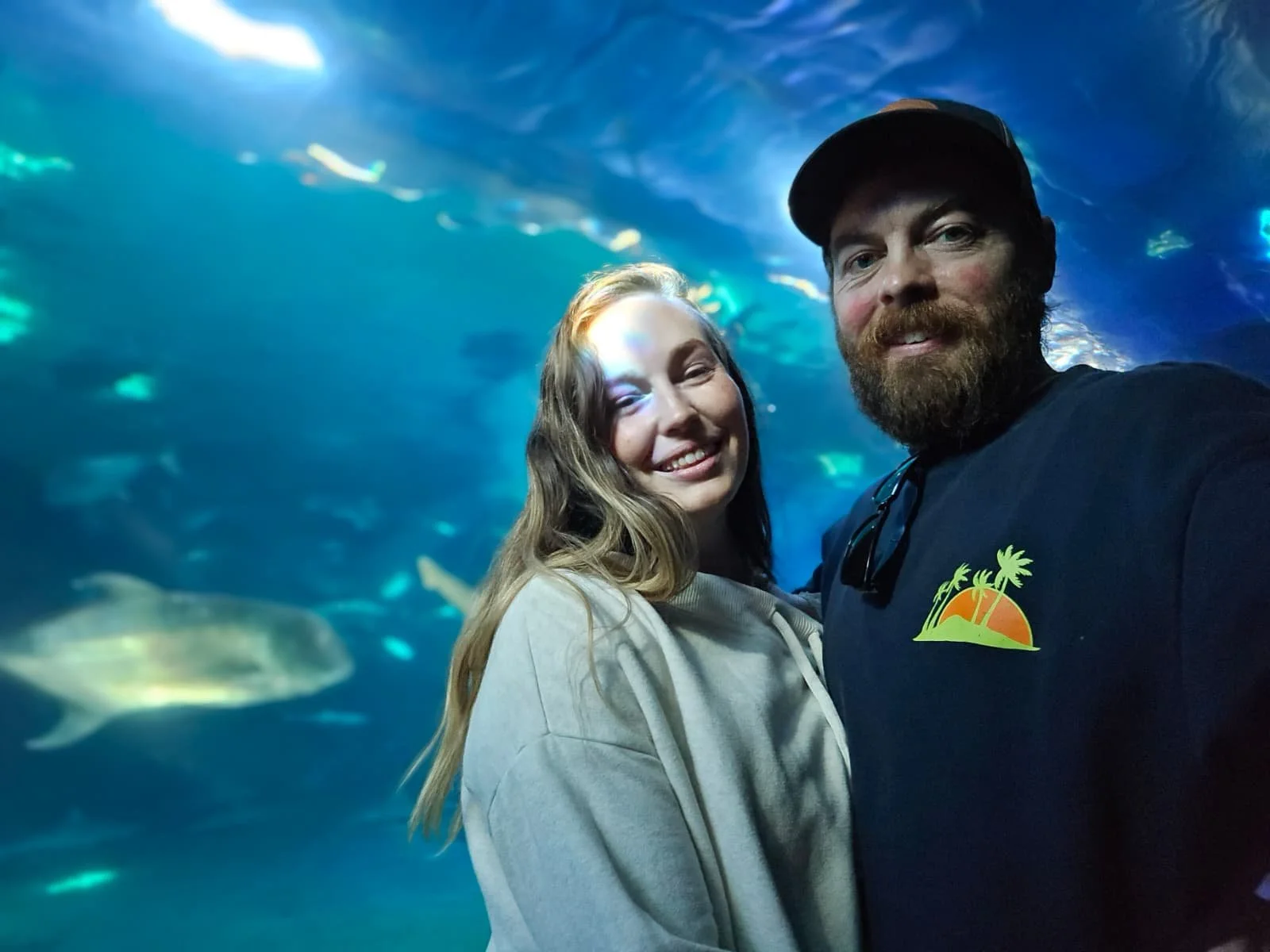 A smiling man and woman, live wedding painter, taking a selfie in front of an aquarium tank on Maui with fish swimming behind them.