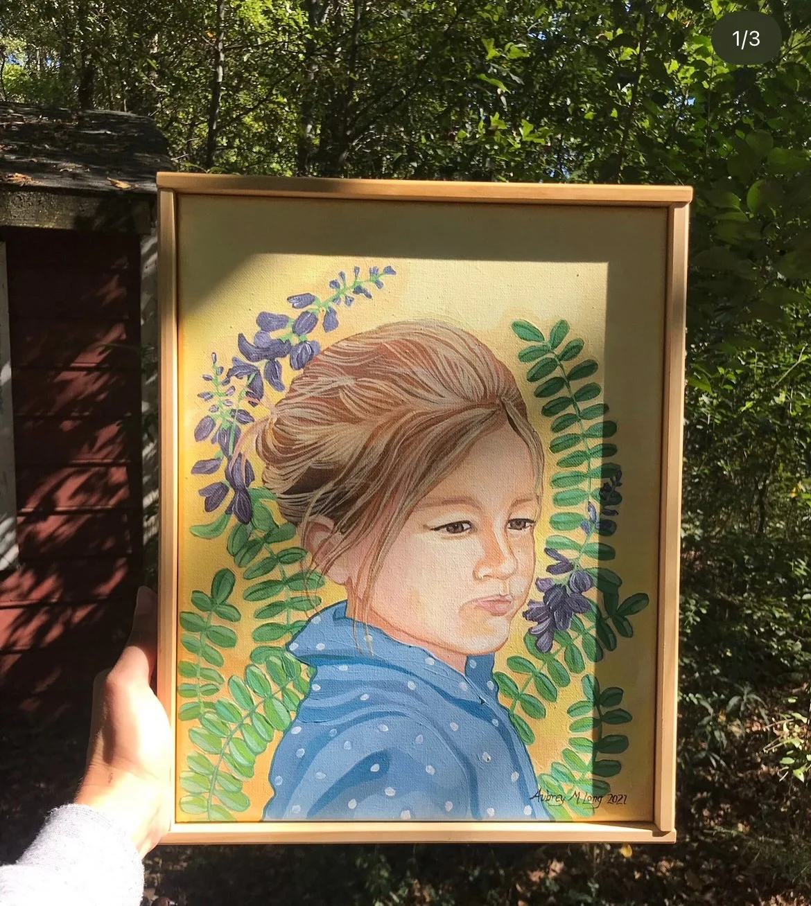 A framed painting of a young girl with light brown hair and a blue polka-dot hoodie, surrounded by green and purple leaves, held outdoors with trees and sunlight in the background.