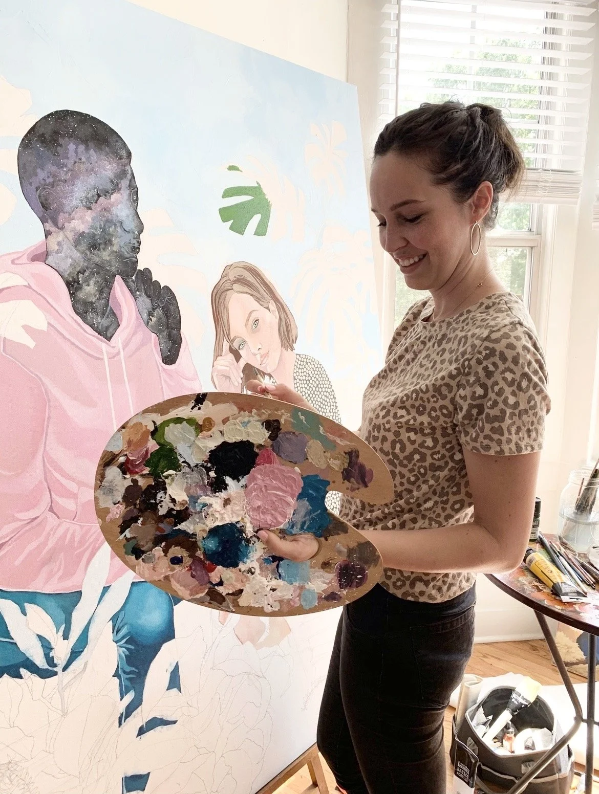Woman smiling while holding a painter's palette with colorful paint, standing in front of a large partially finished painting of two women with leafy background.
