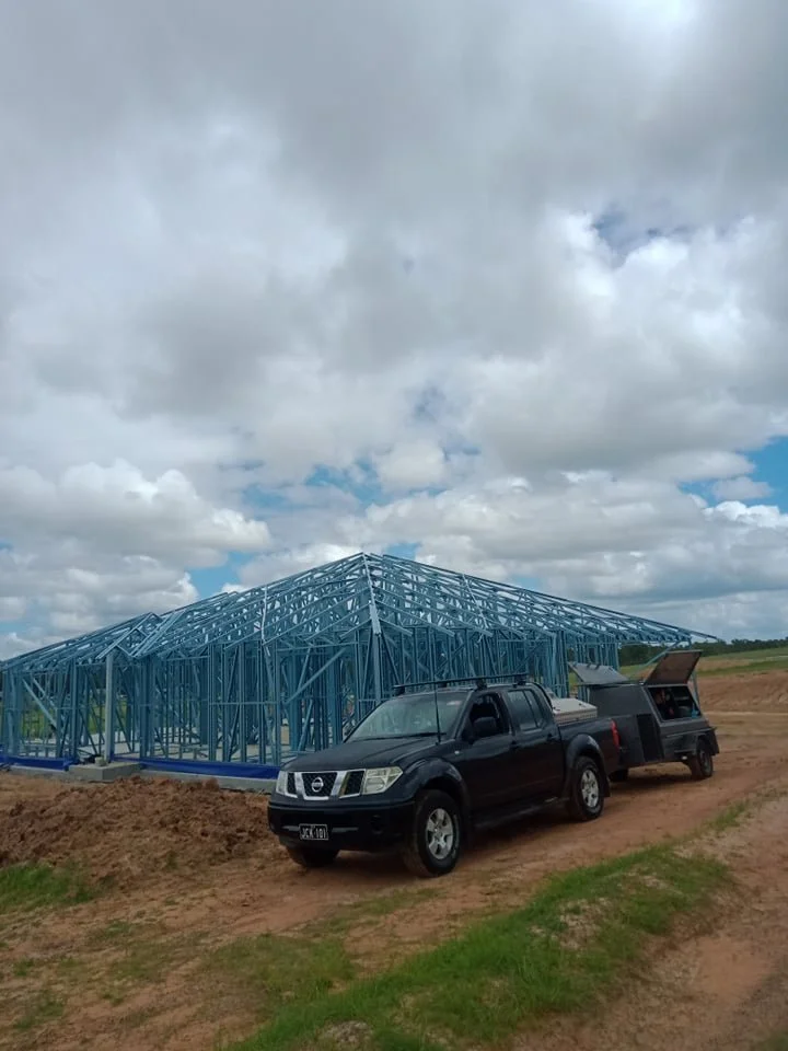 Blue metal framework of a building under construction on a dirt field with a black pickup truck and trailer nearby, cloudy sky overhead.
