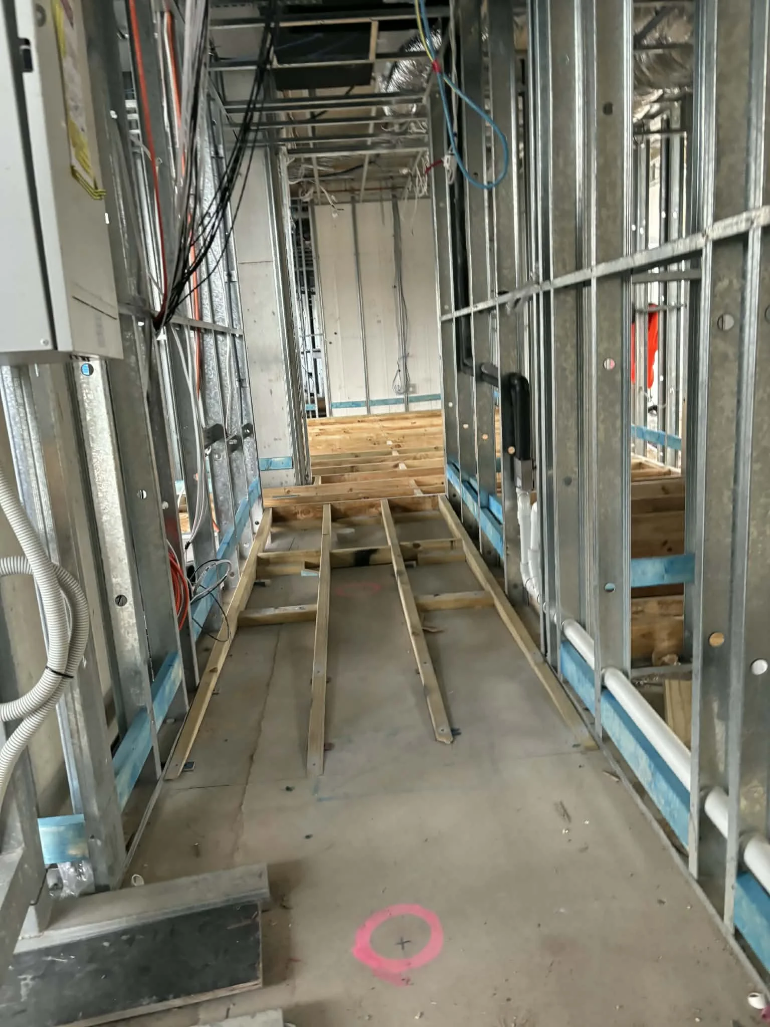 Construction site inside a building with exposed metal framing, electrical wiring, and unfinished flooring. Wooden floor joists are being installed.