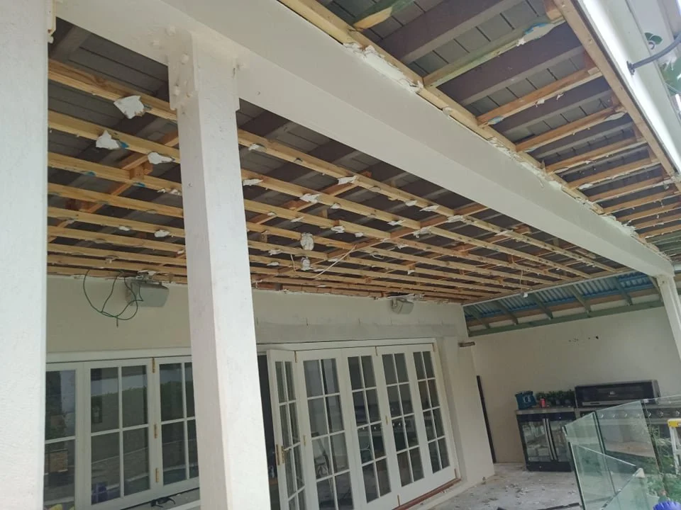 Construction site showing an unfinished balcony ceiling with exposed wooden beams and insulation, with glass sliding doors and a grill in the background.