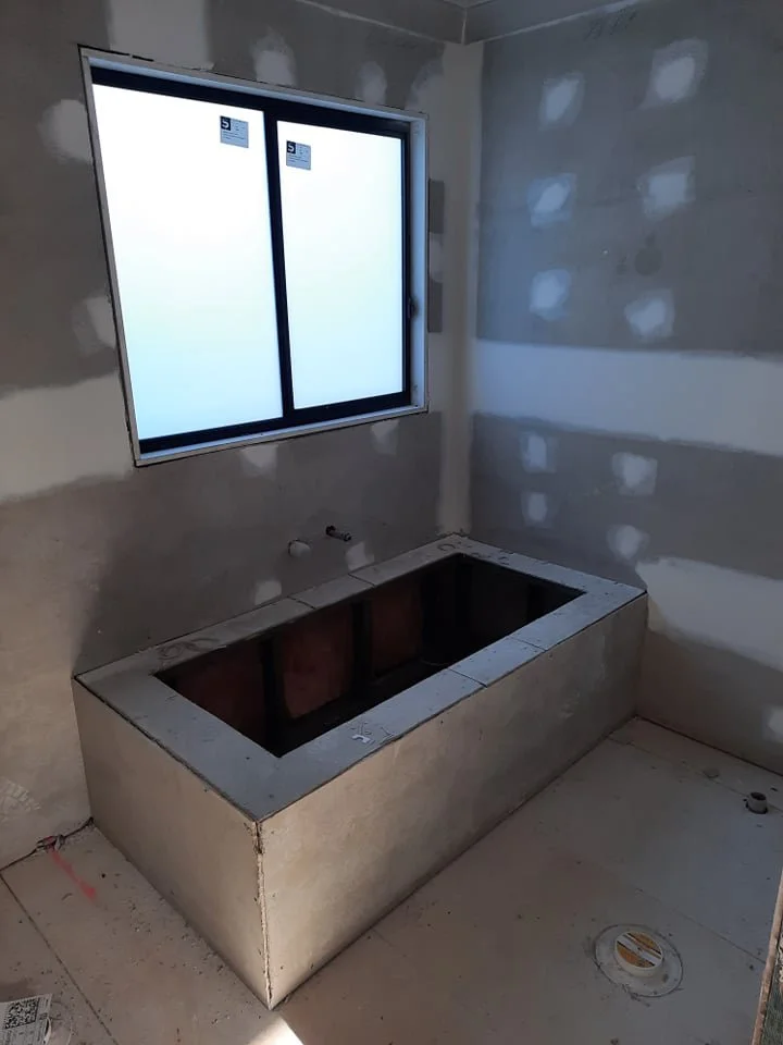 Unfinished bathroom with a large rectangular bathtub frame made of concrete or cement, installed below a window in a room with drywall walls that are partially finished.