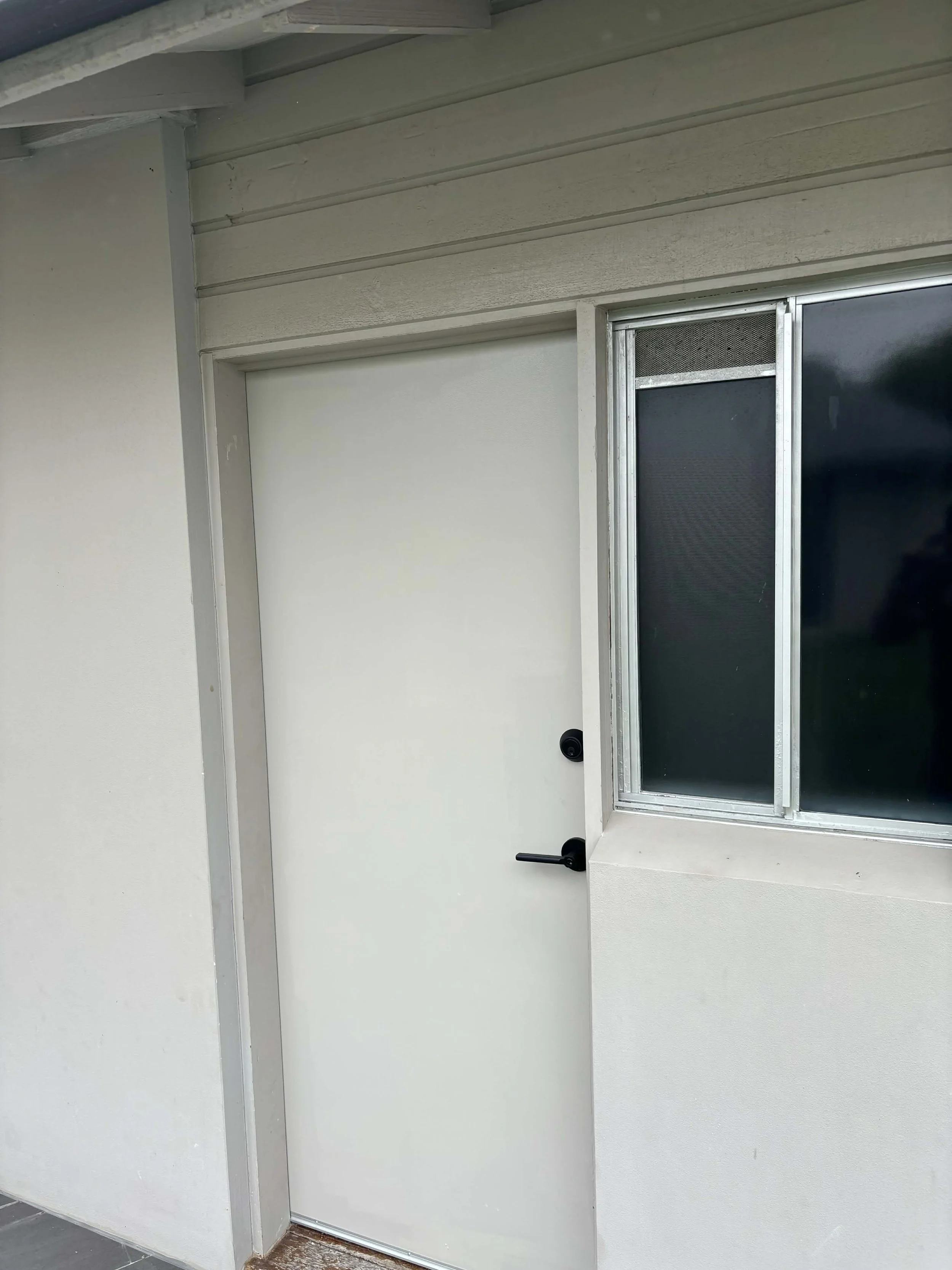 White door with black handle next to a window with black screens on a beige wall.