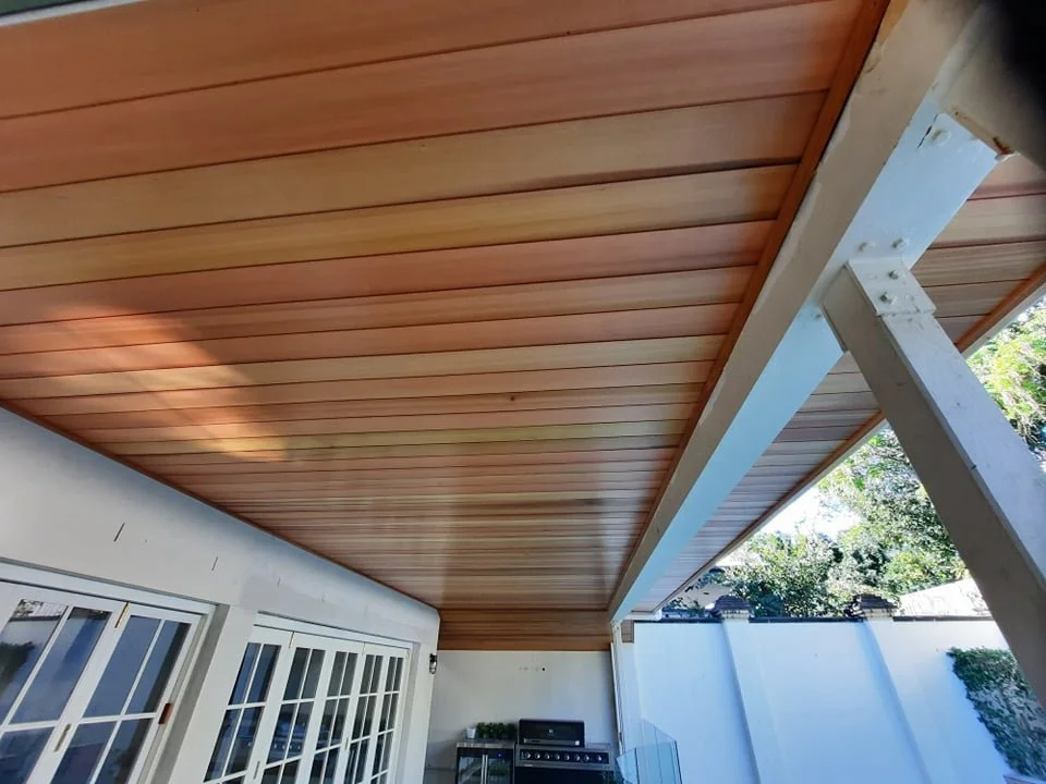 A wooden patio ceiling with a white support beam and a glimpse of an outdoor grill in a backyard.
