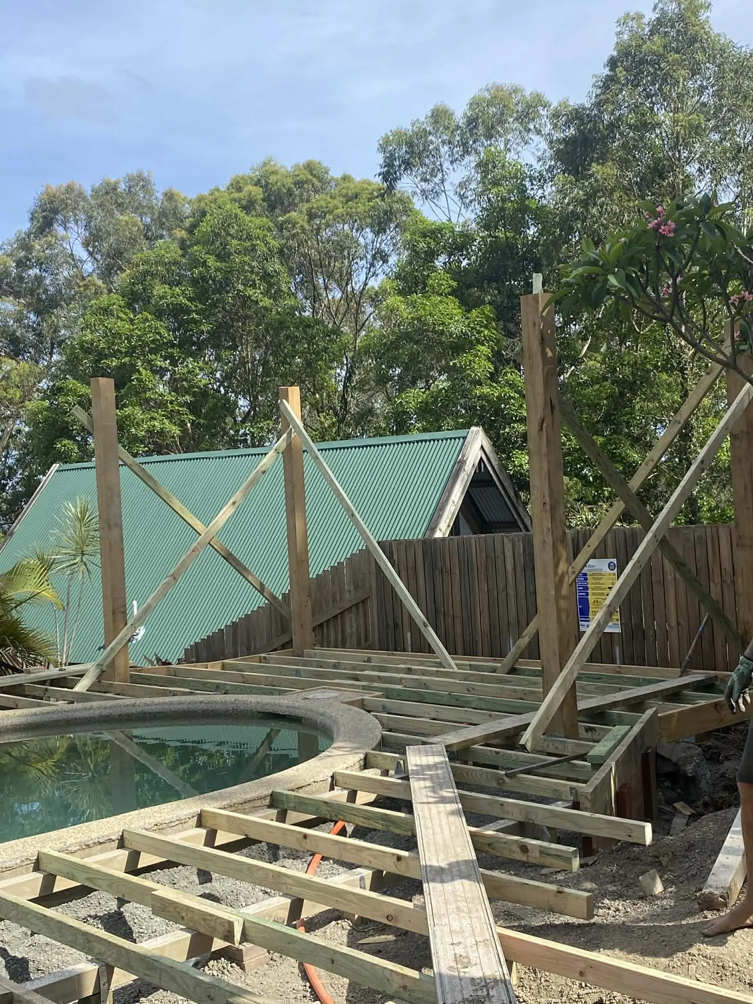 Construction site with wooden framework in progress near a pool, surrounded by trees and a wooden fence.