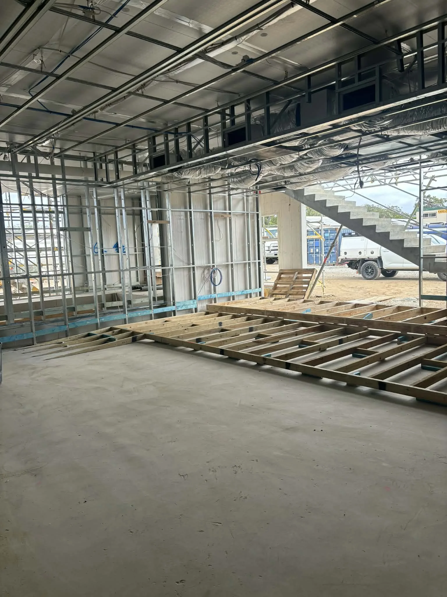 Construction site showing an unfinished building with exposed framing, electrical wiring, and a staircase leading to an upper level. The floor is concrete, and outside the open wall, construction vehicles are visible.