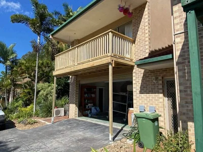 Two-story brick house with a wooden balcony above a garage, surrounded by palm trees and greenery, with a paved driveway and a green trash bin outside.