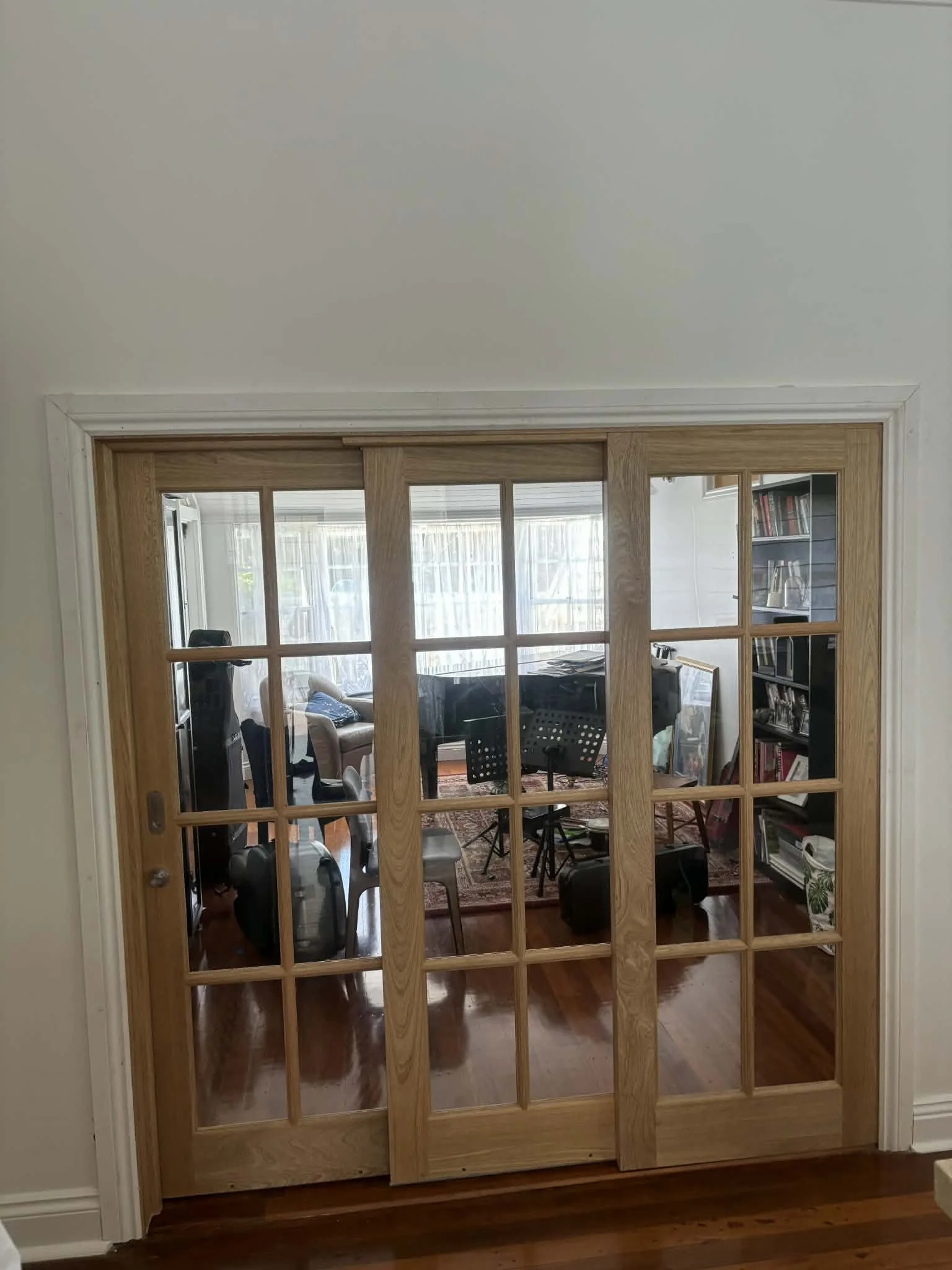 A wooden sliding door with glass panes separates two rooms, with the front room painted white and the room behind filled with furniture, bookshelves, and musical equipment.