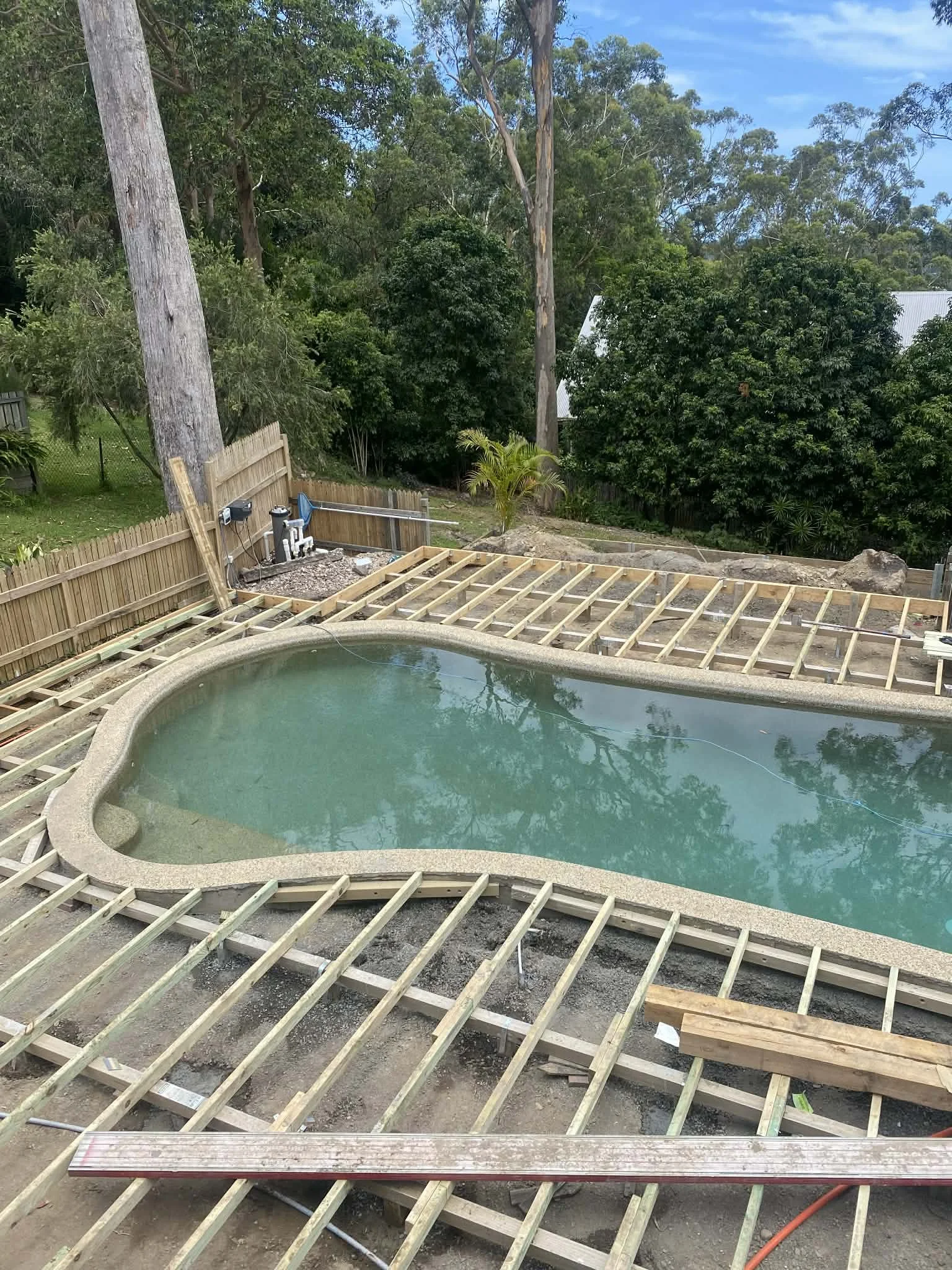 A backyard with a small kidney-shaped swimming pool under construction, surrounded by wooden framing, with trees and greenery in the background.