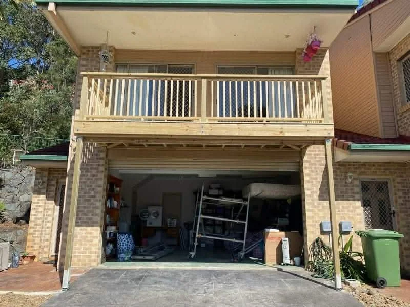 Front view of a two-story brick house with a partially open garage on the ground level, storing various items, and a wooden balcony on the upper floor with a railing, flower decorations, and a green trash bin outside.