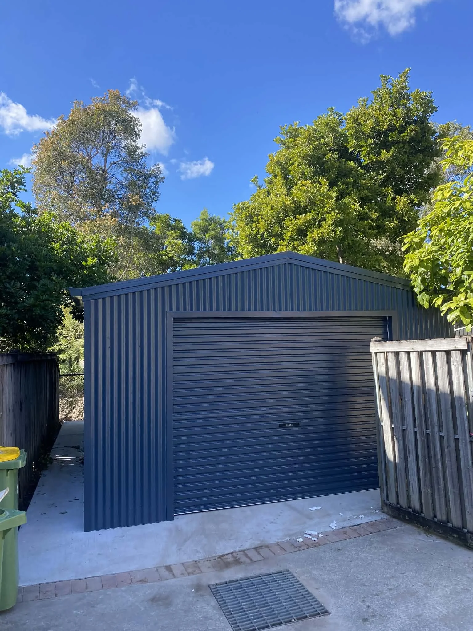 A blue metal garage with a roll-up door, surrounded by a wooden fence on the right and green trees in the background, under a bright blue sky with a few clouds.