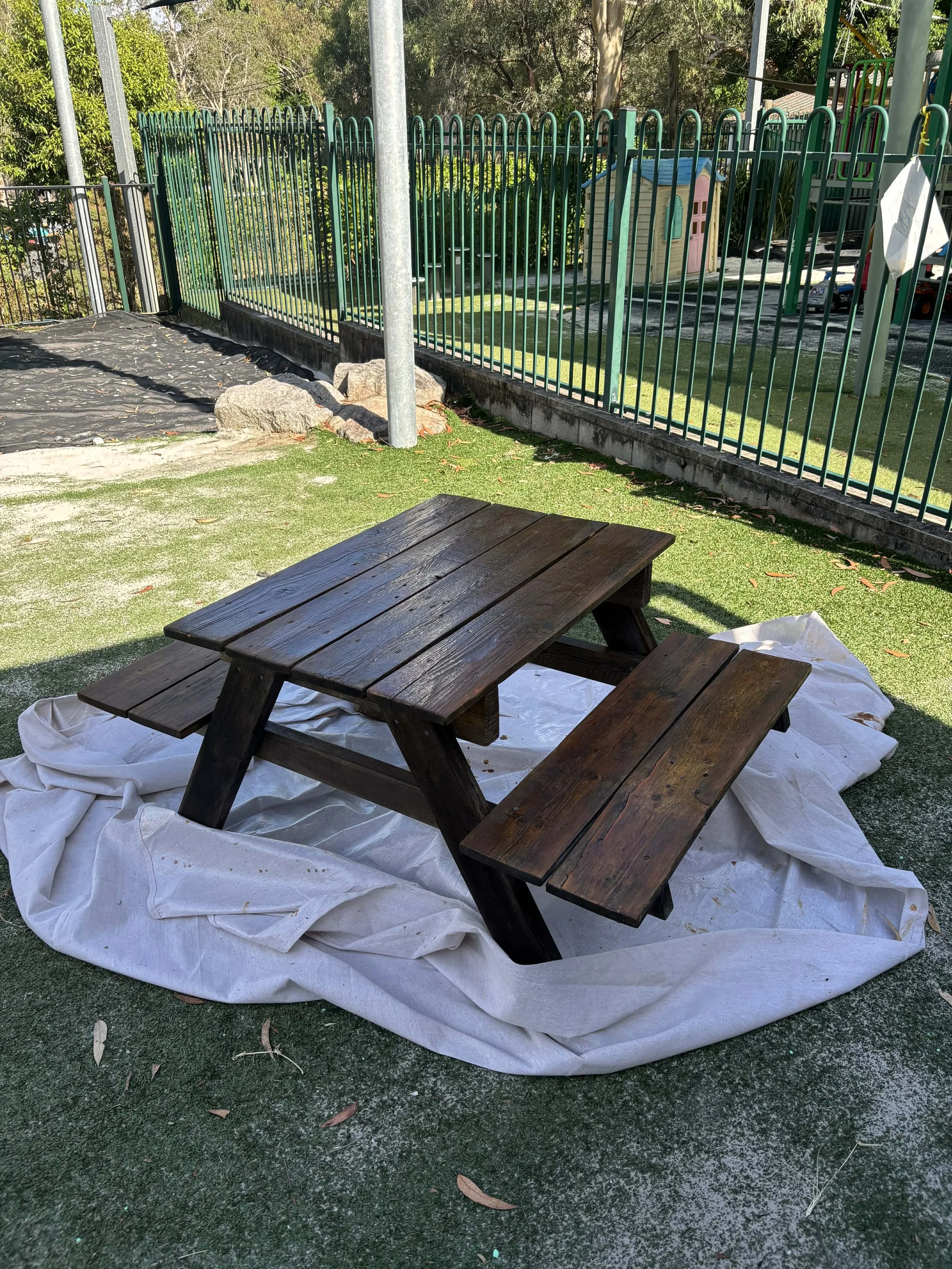 A wooden picnic table with attached benches on a white cloth cover, placed on a grassy area surrounded by a fence, with playground equipment and trees in the background.