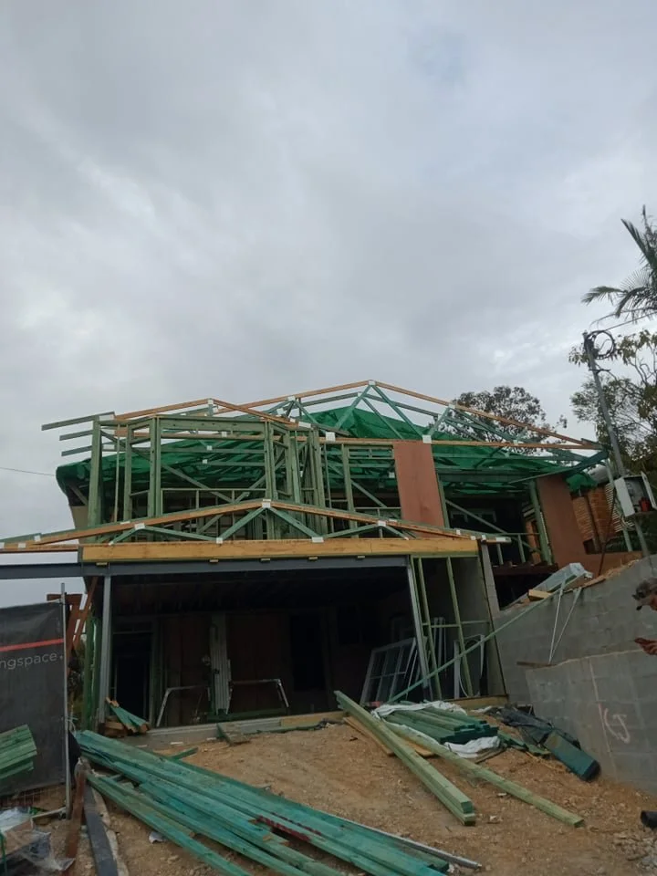 Construction site of a house with wooden framing and green roofing under cloudy sky.