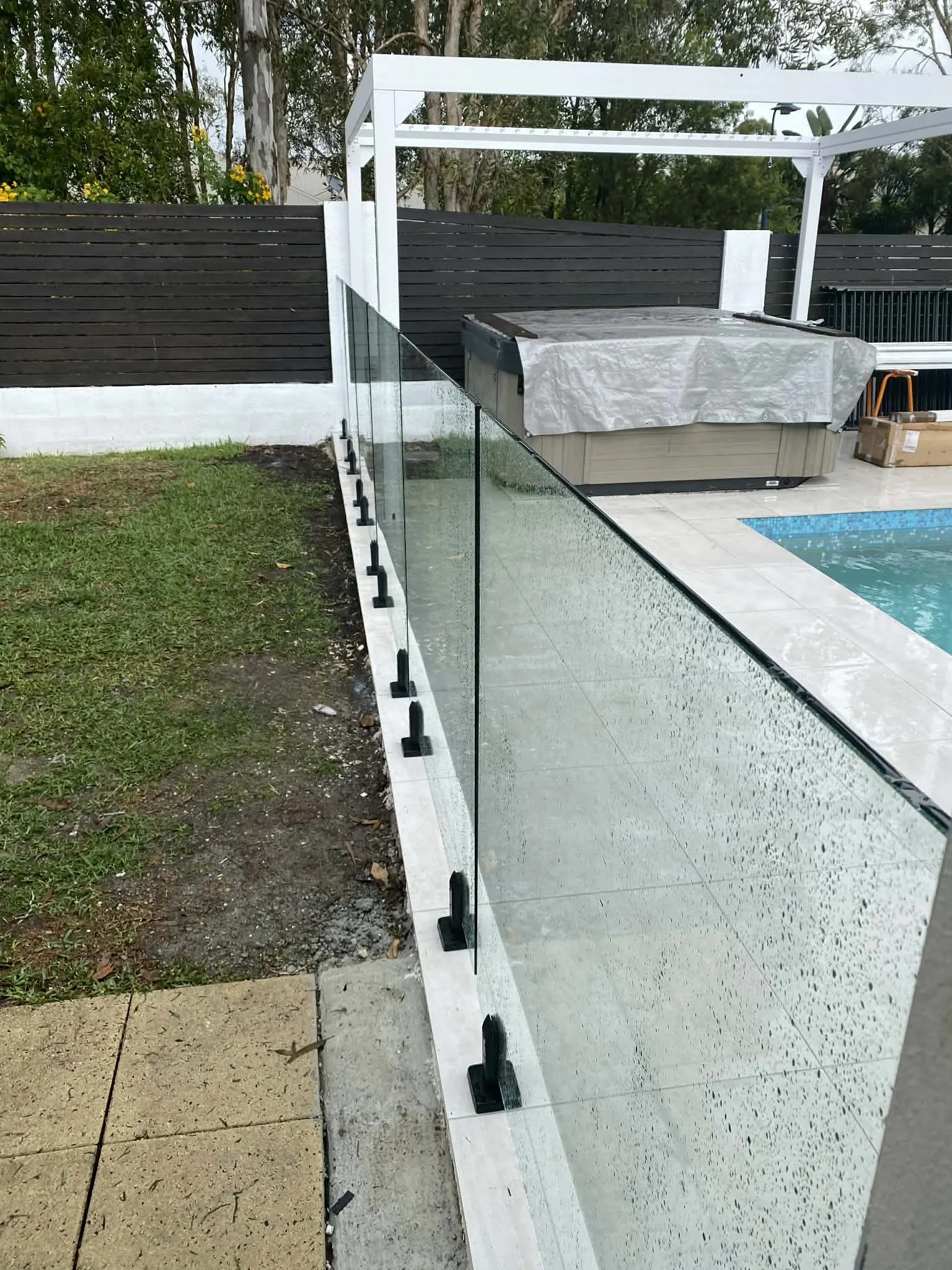 A newly installed glass pool fence along the edge of a swimming pool, with a concrete patio and a hot tub covered with a tarp in the background.