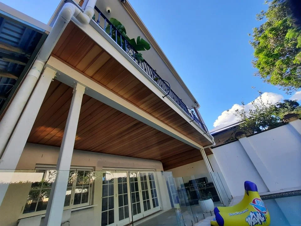 A two-story house with a balcony and a pool in the backyard, with blue sky and trees surrounding the area.