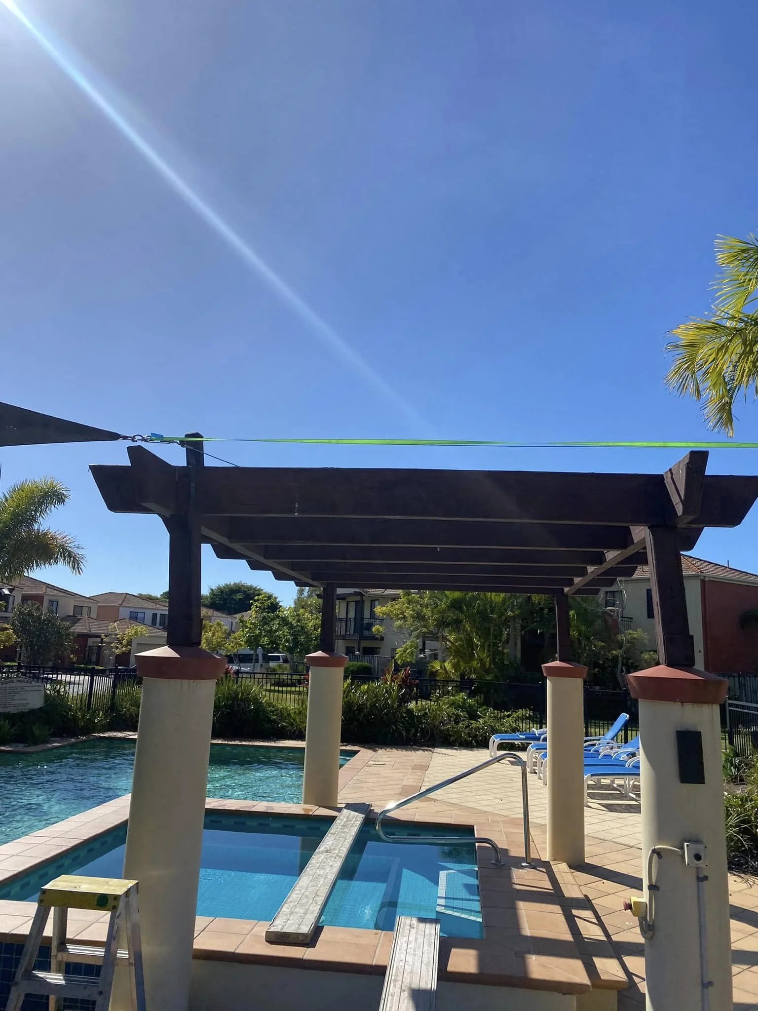 Swimming pool with a shaded lounge area, lounge chairs, and lush greenery in the background under a clear blue sky.